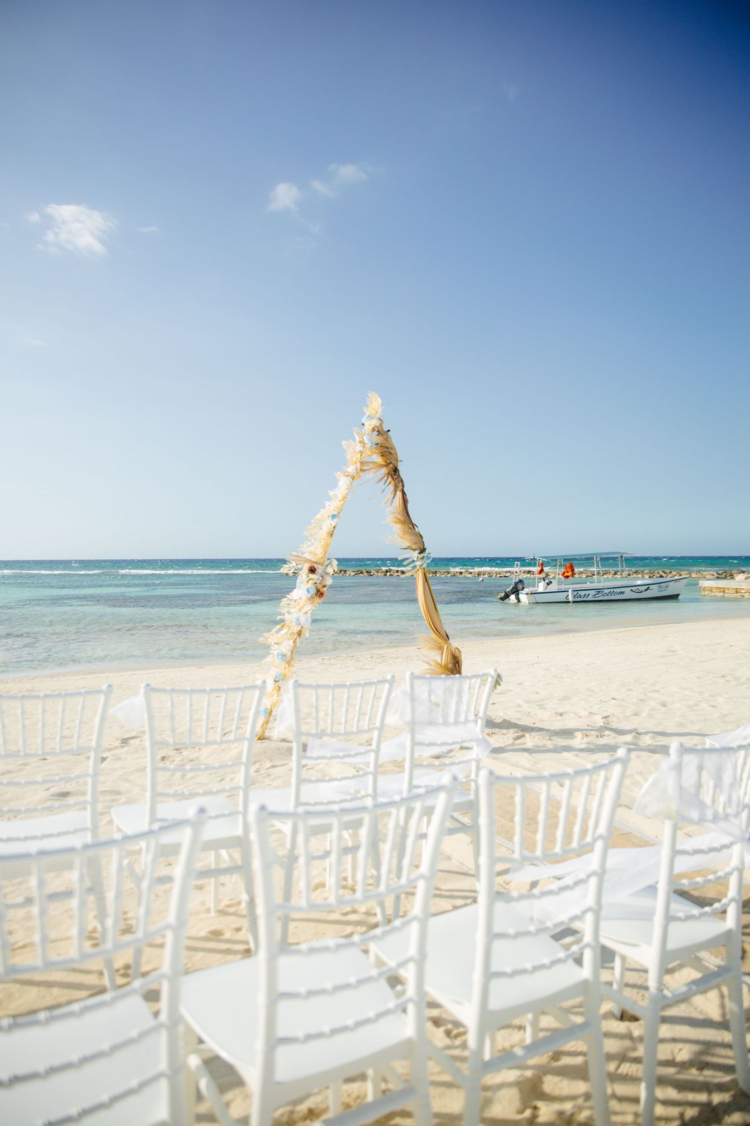 White chiavari chairs line the aisle toward a rustic triangular arch on a pristine Caribbean beach wedding setup.