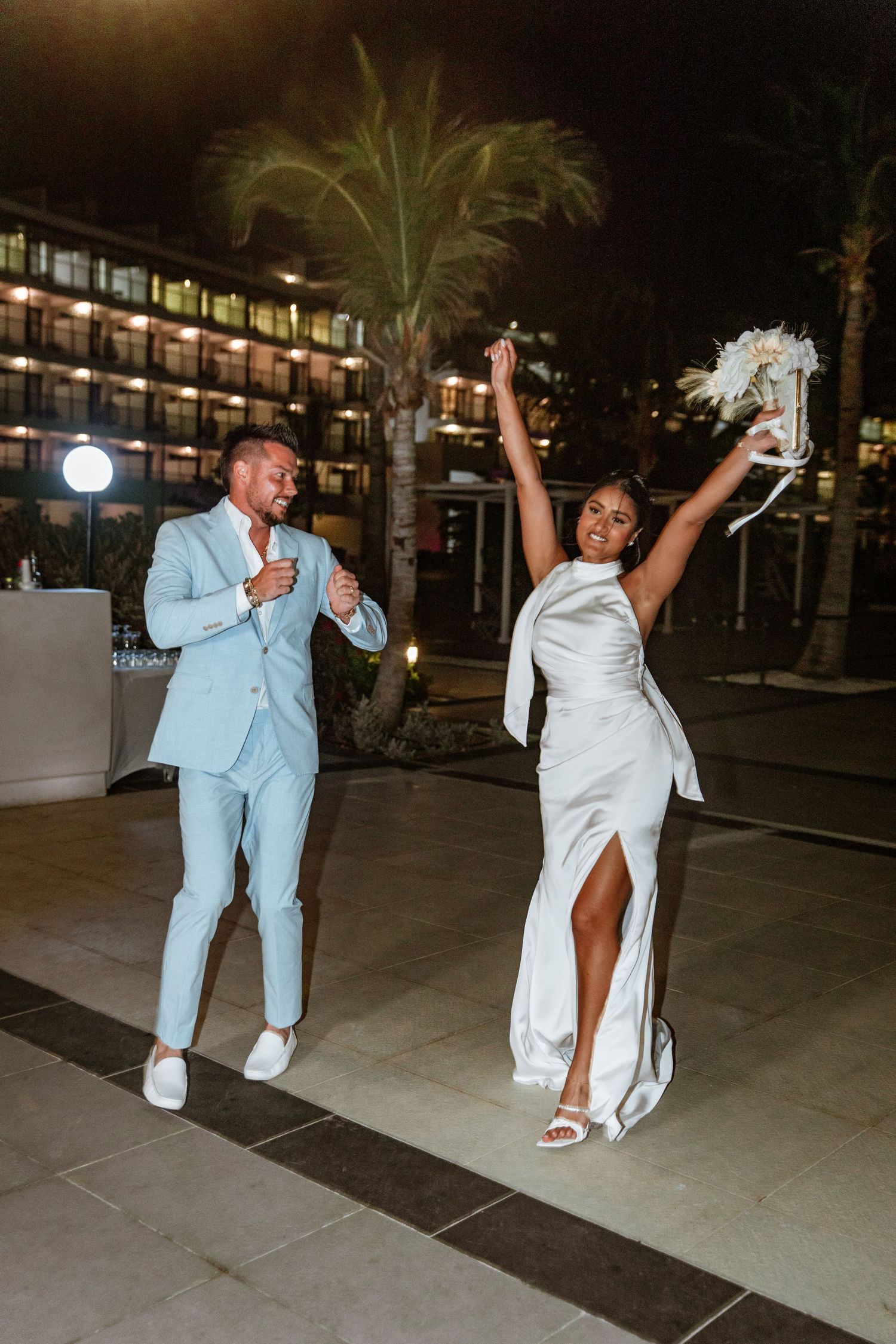 Joyful newlyweds dancing and celebrating outside hotel at night bride raising bouquet.