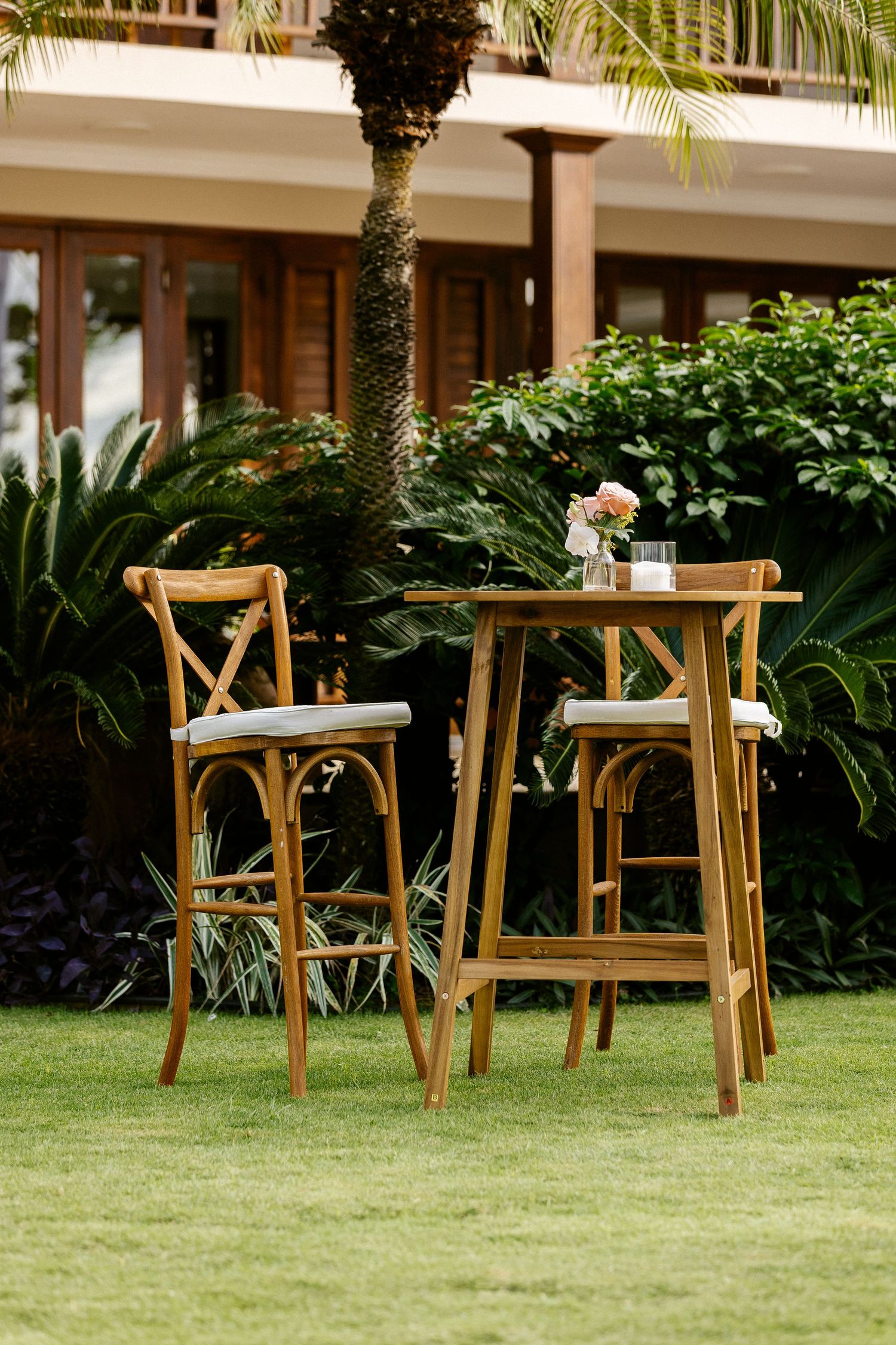 Wooden bar-height bistro table with two cross-back stools on green lawn, tropical garden and resort building backdrop.