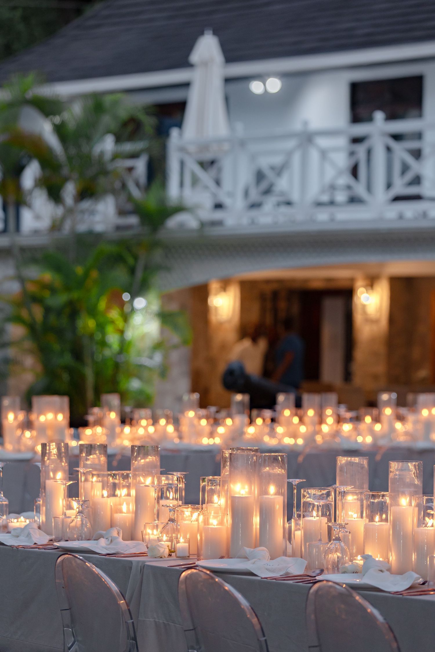 Candlelit outdoor wedding reception tables glowing at dusk with white pillar candles and colonial-style venue in background.
