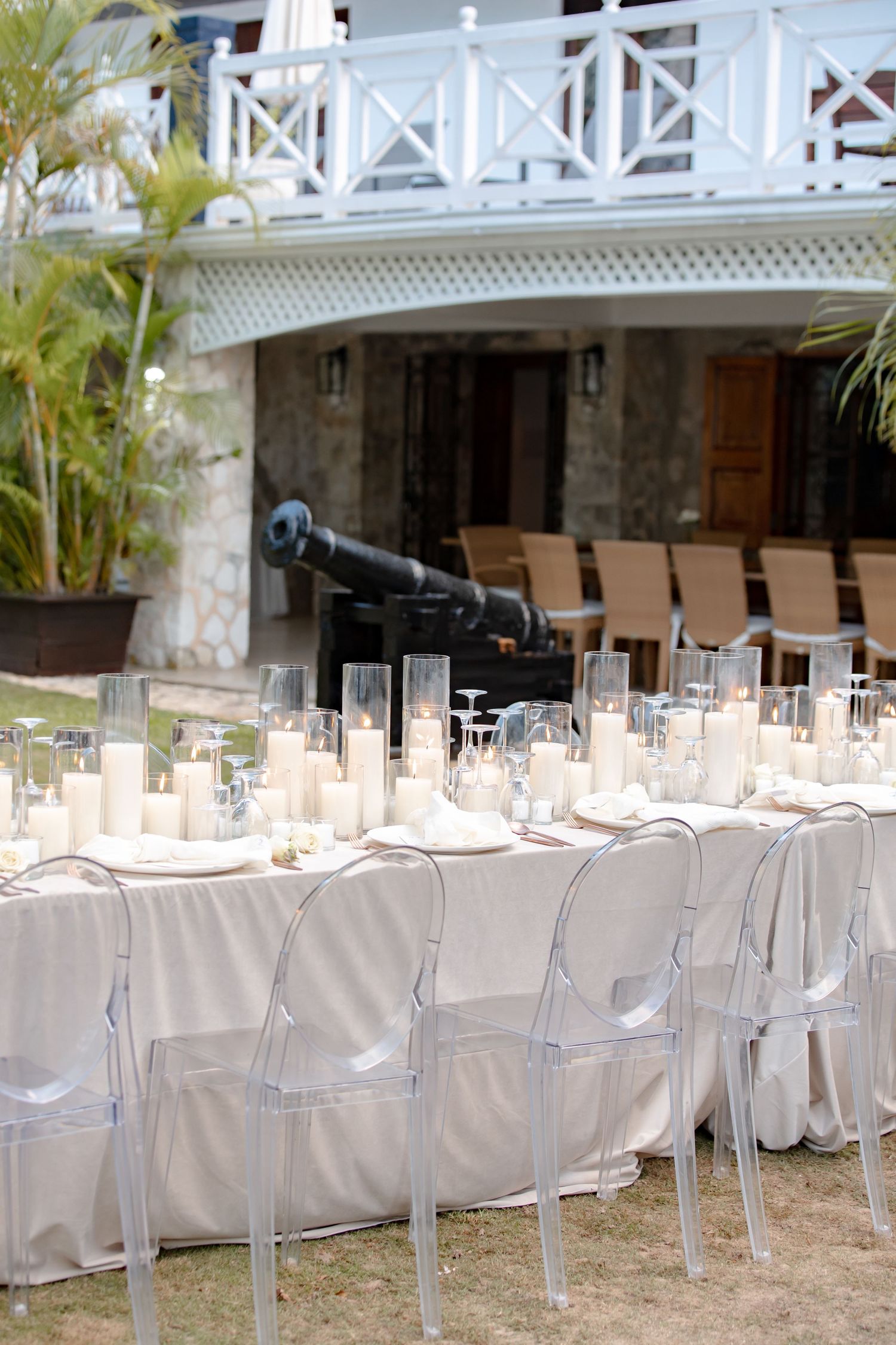 Elegant outdoor wedding reception table with ghost chairs, white candles, and a historic cannon centerpiece near a tropical villa.