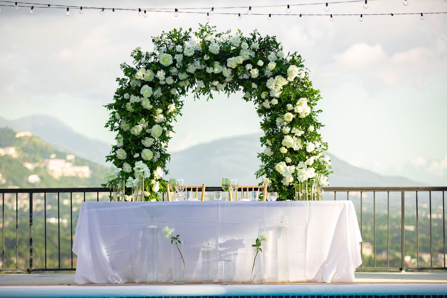 Elegant outdoor wedding arch adorned with white flowers and greenery over a draped sweetheart table with mountain views.