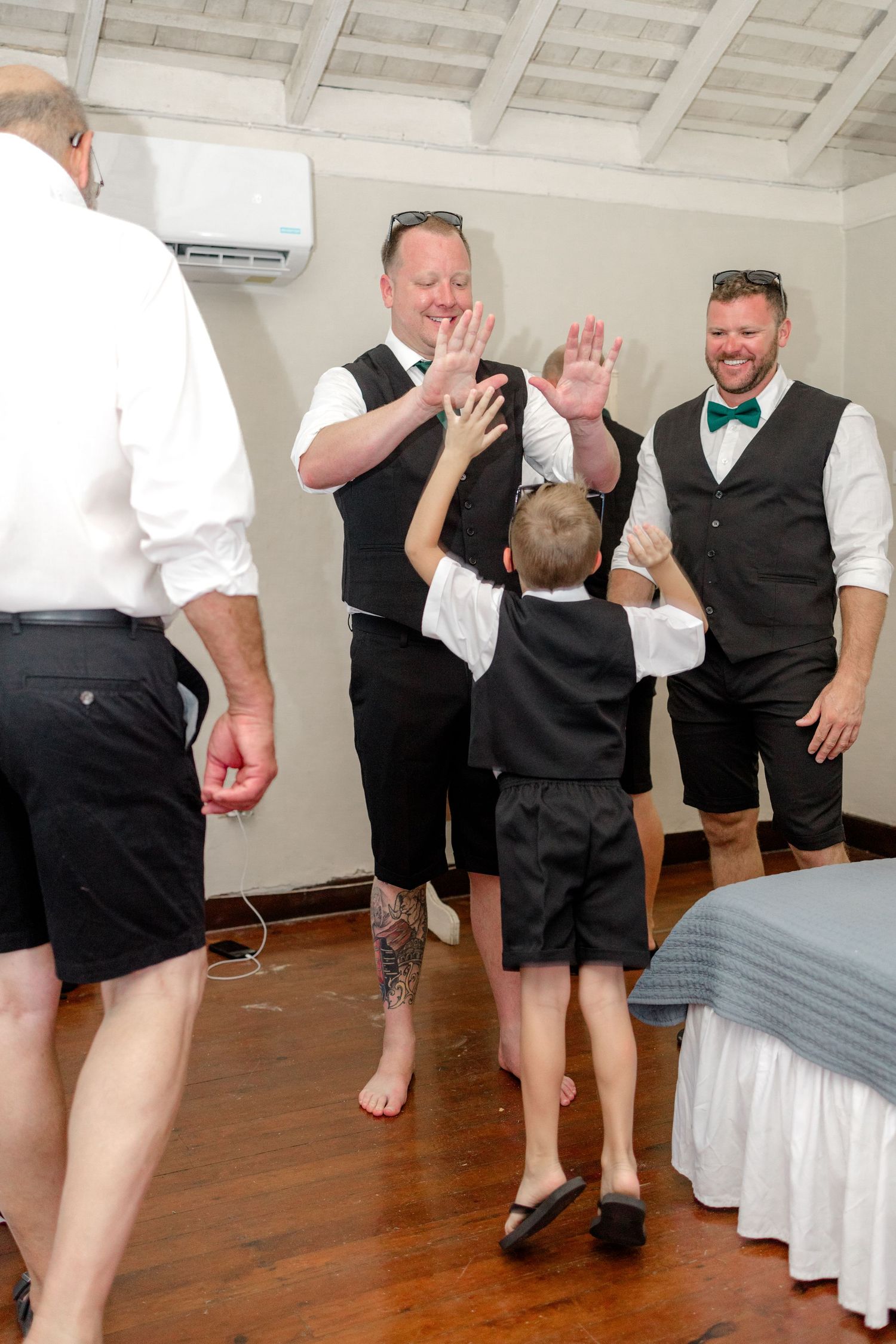 Groomsmen in black vests high-five an excited young boy in a wedding venue room.