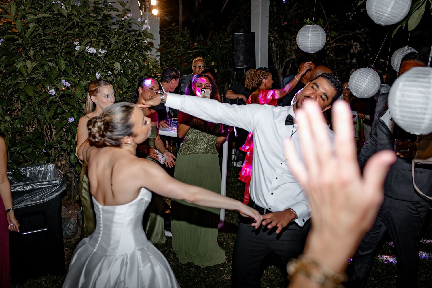 Bride dances energetically with a guest in a white suit at a lively outdoor wedding reception at night.