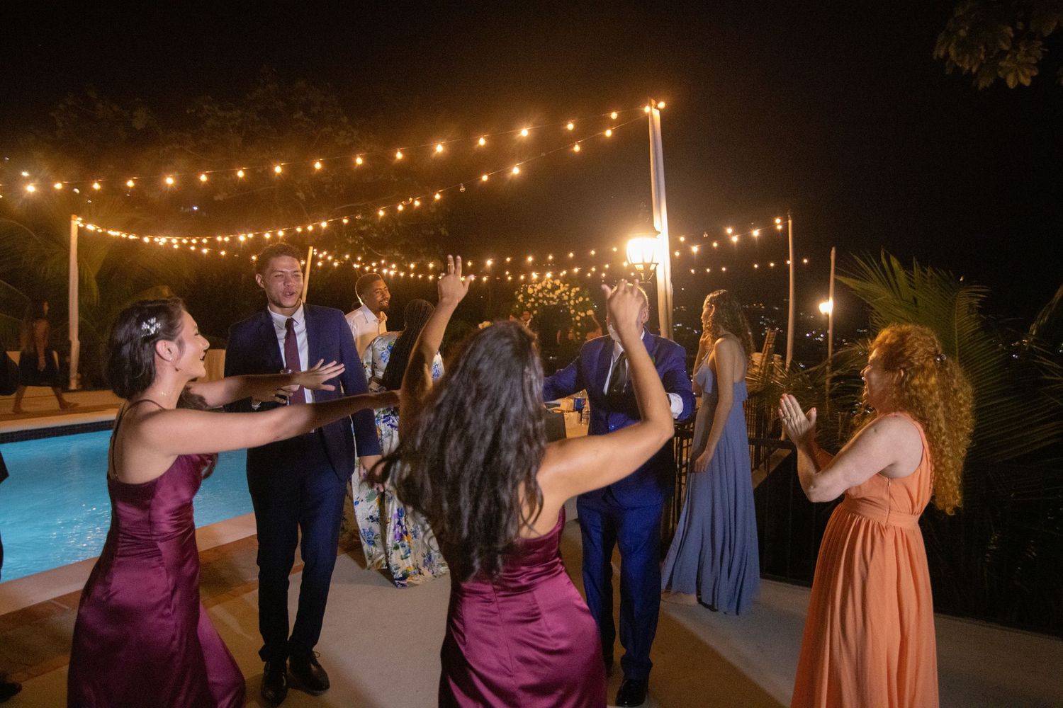 Wedding guests dancing outdoors at night under string lights near a pool.