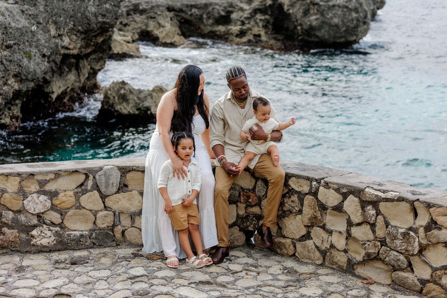 Family of four posing on a stone wall by turquoise ocean waters near rocky cliffs in a tropical destination.