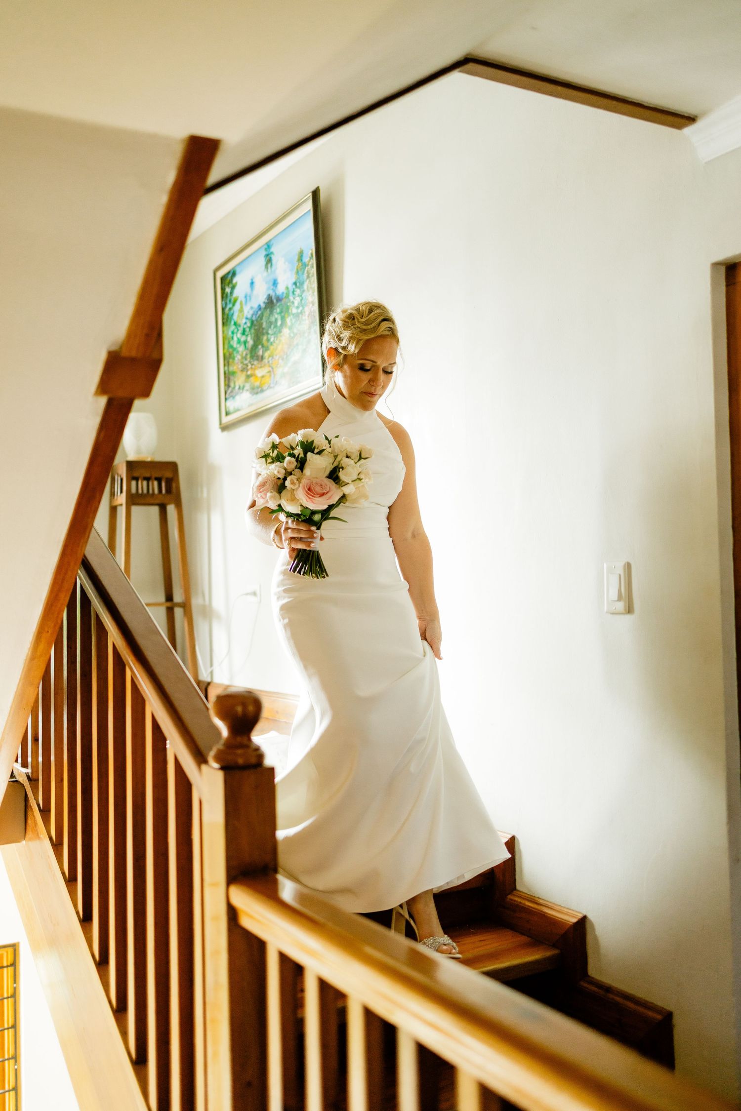Bride in white halter gown holding pink bouquet descends wooden staircase in bright home interior.