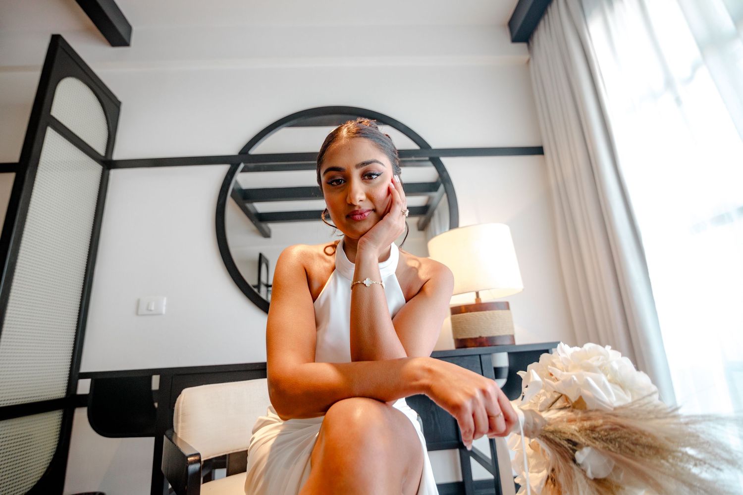 A smiling woman in a white dress sits elegantly in a bright, modern hotel room with decorative round mirror.
