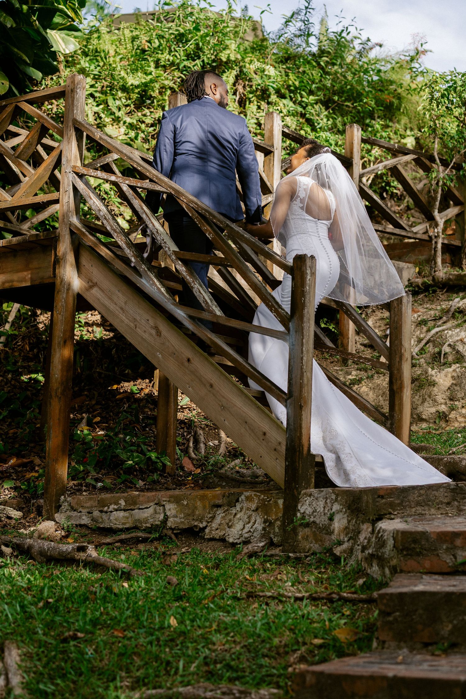 Bride and groom walking up rustic wooden stairs surrounded by lush greenery on their wedding day.