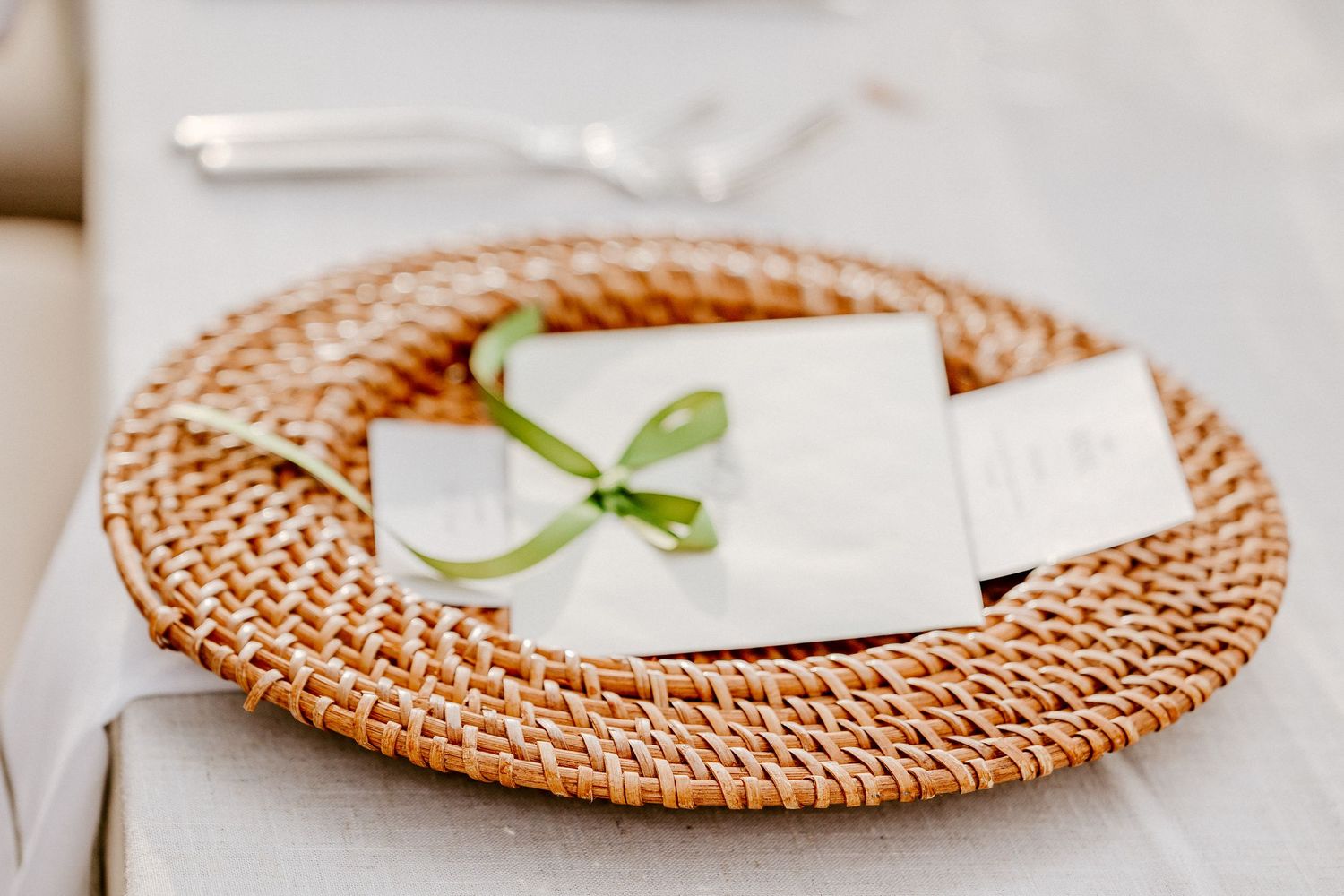 Woven rattan charger plate with white envelope tied with green ribbon on elegant table setting.