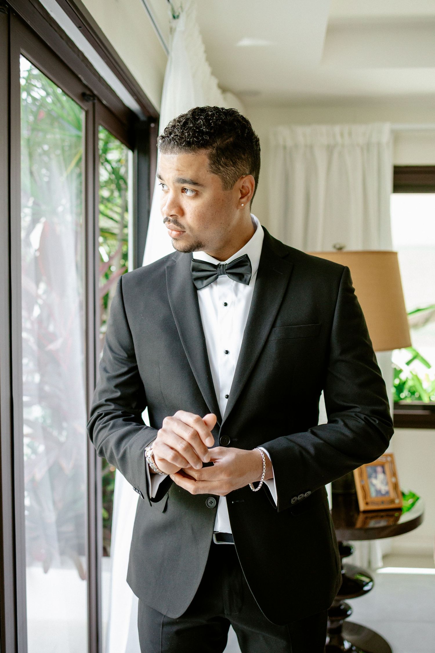 Elegant groom in black tuxedo with bow tie standing by window adjusting cufflinks on wedding day.