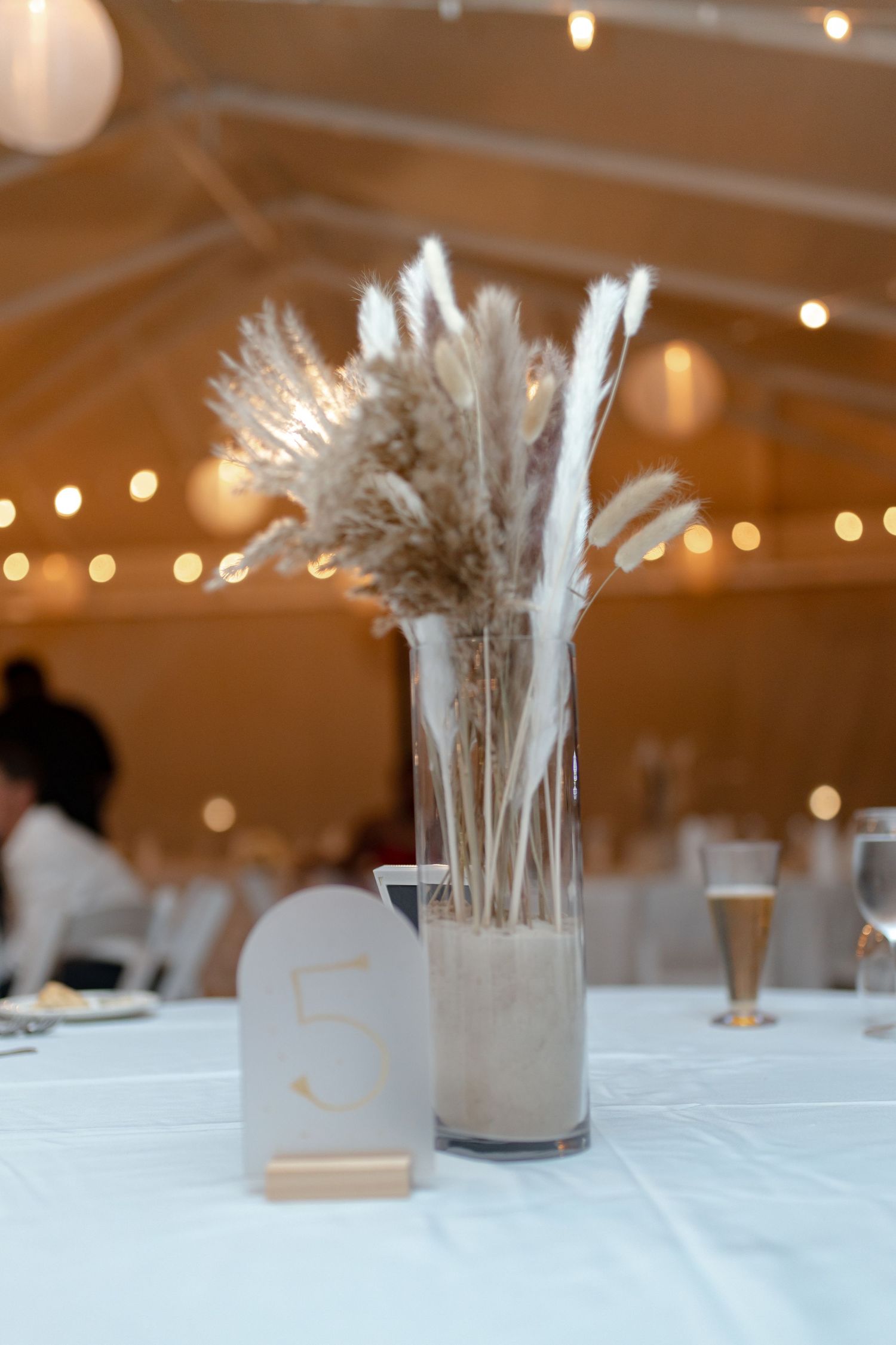 Elegant boho wedding centerpiece with pampas grass in glass vase on white reception table.