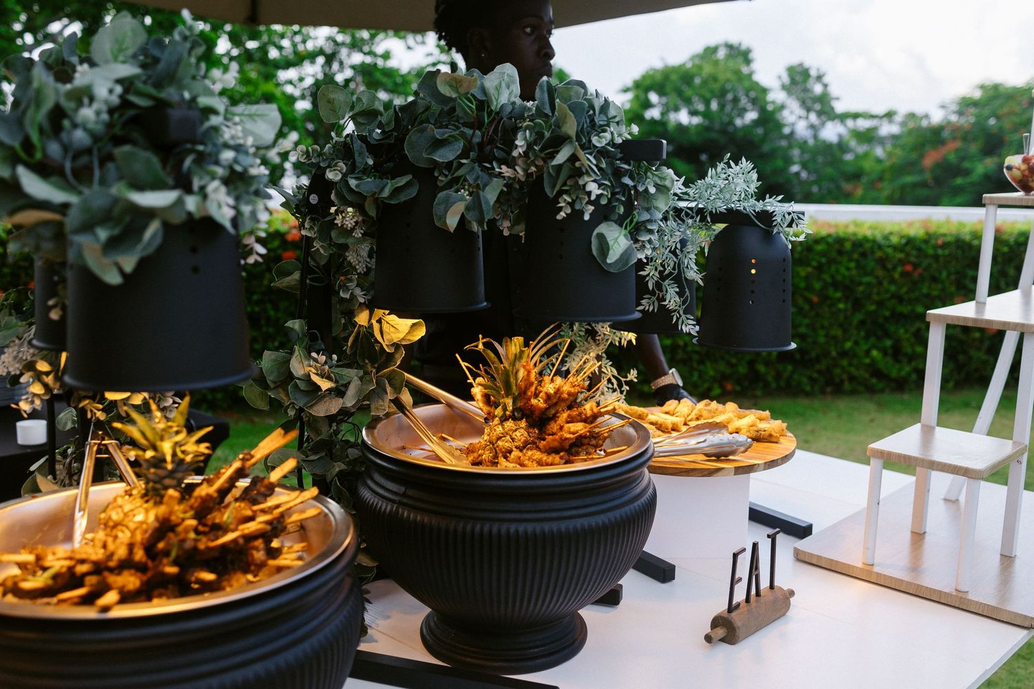 Elegant outdoor wedding buffet display with tropical greenery and golden food arrangements.