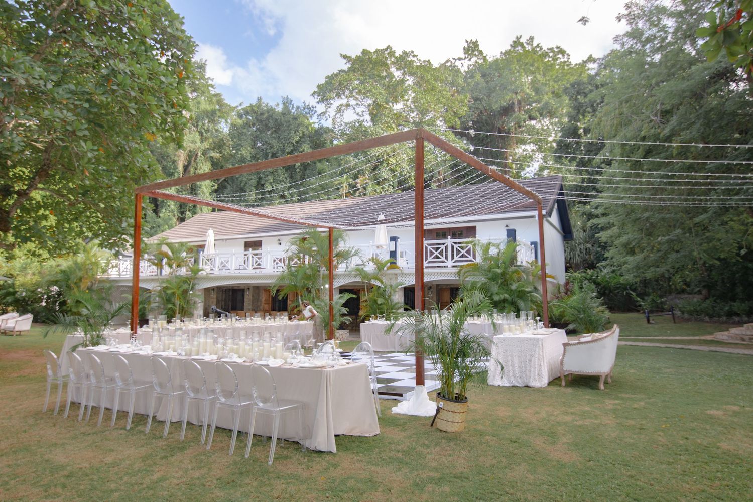 Elegant outdoor wedding reception setup with white chairs and long tables under a wooden pergola on a lush green lawn.
