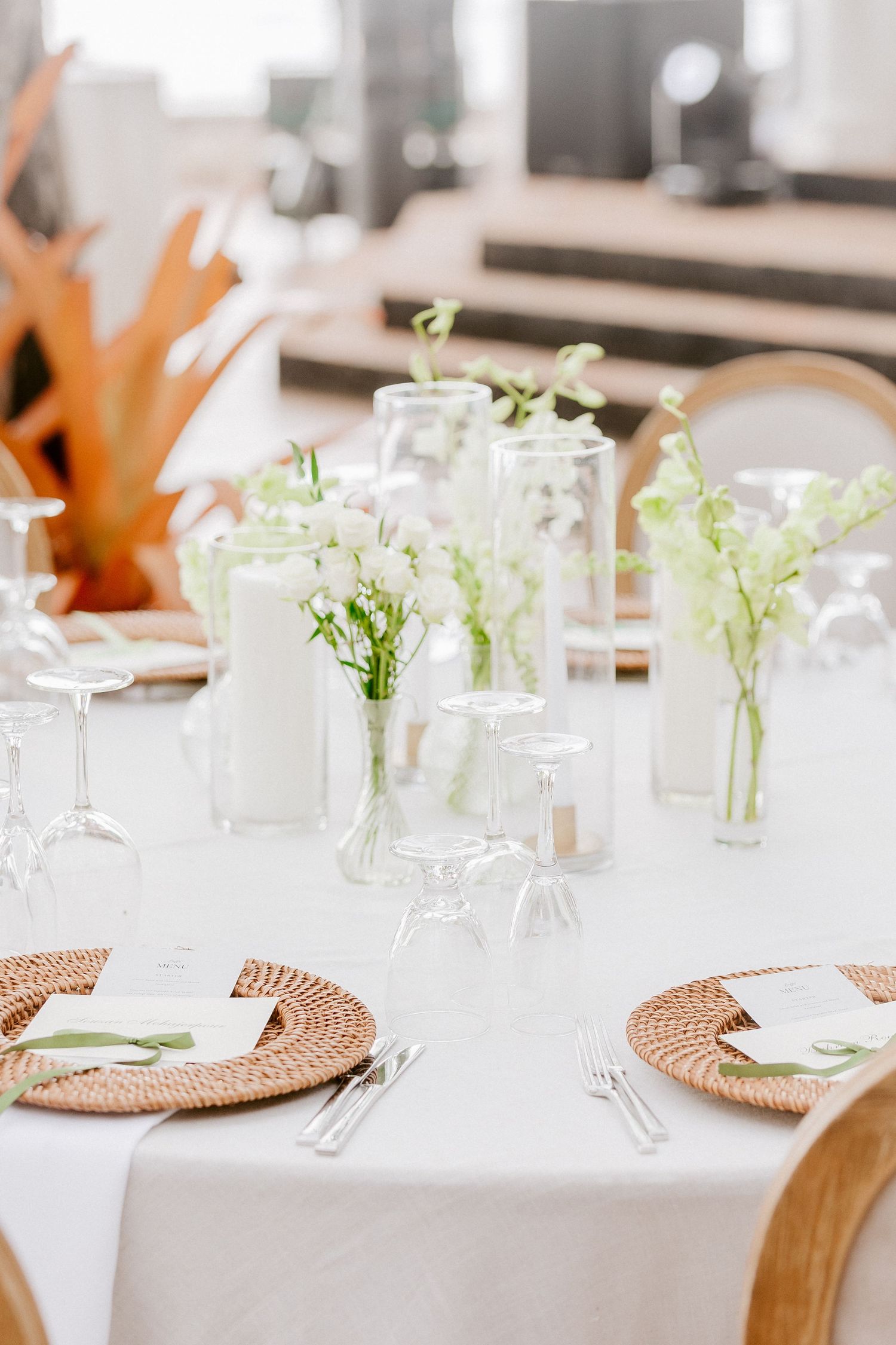 Elegant wedding reception table with wicker chargers, white florals in glass vases, and crystal stemware on white linen.