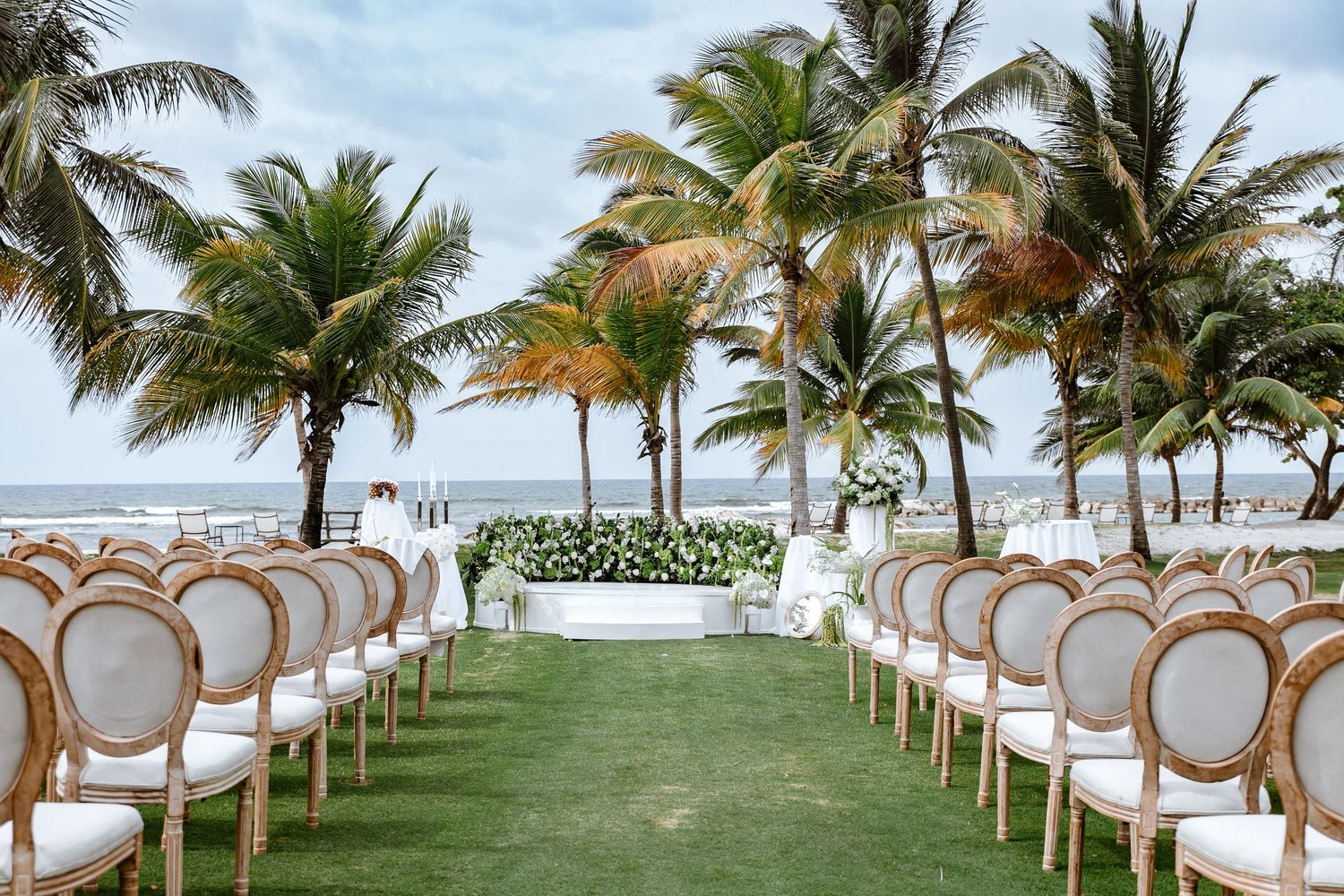 Elegant outdoor beach wedding ceremony setup with white chairs, palm trees, and ocean view on lush green lawn.