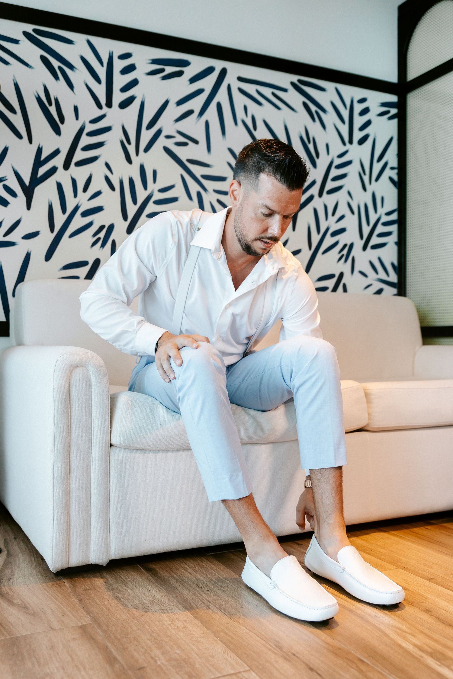 Groom in white shirt and light blue pants sitting on white sofa putting on white loafers before wedding.