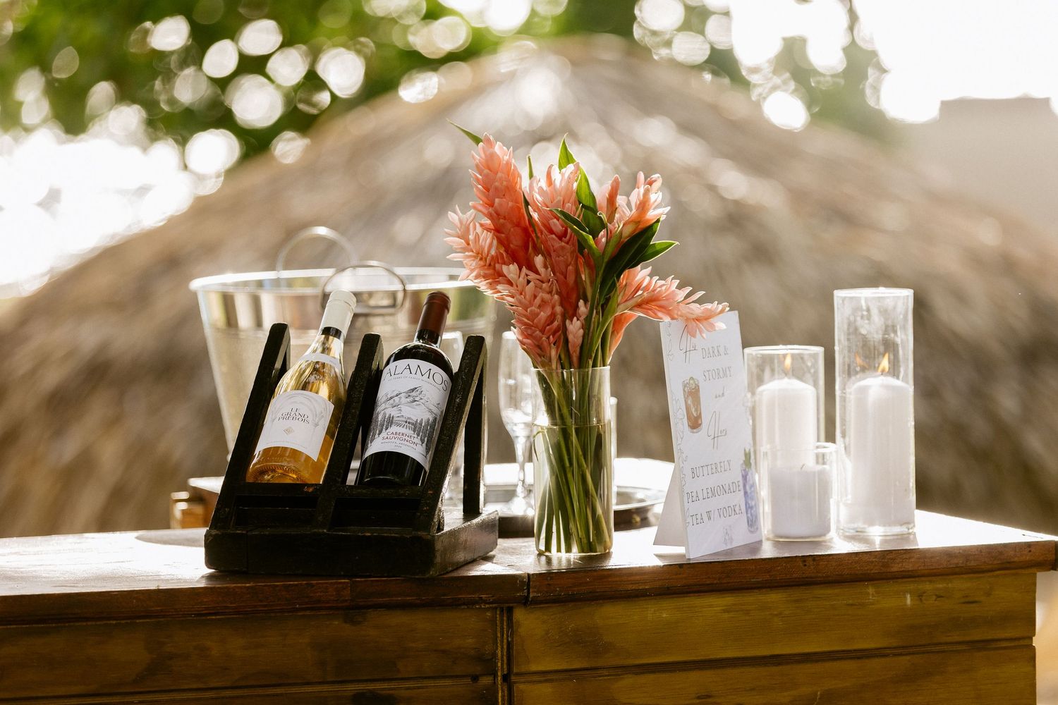 Wooden table with two wine bottles, pink floral arrangement, white candles, and a thatched roof backdrop at an outdoor event.