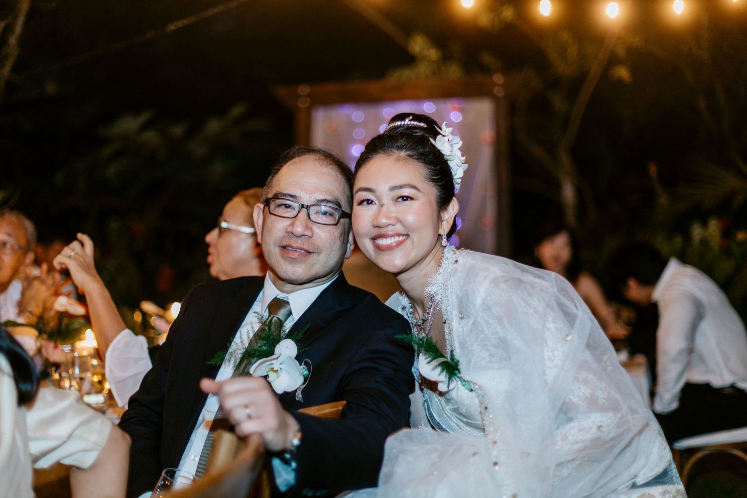Joyful bride in white lace gown poses with suited guest at outdoor evening wedding reception.