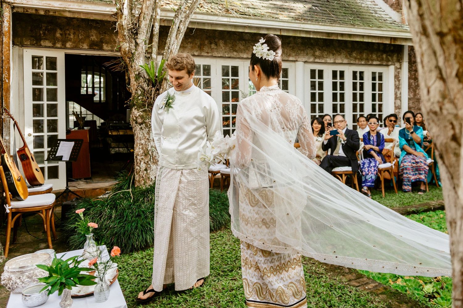 Bride and groom in traditional white and gold wedding attire holding hands during an outdoor ceremony.