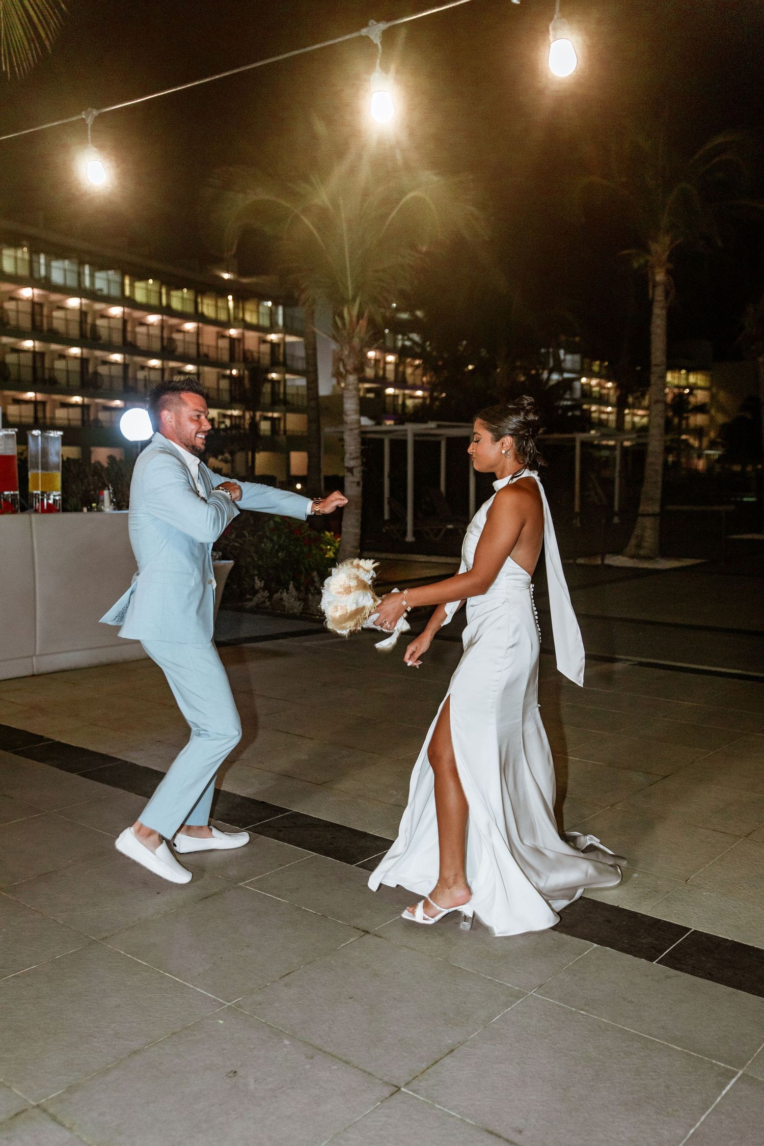 Bride and groom dancing playfully under string lights at outdoor nighttime destination wedding reception.