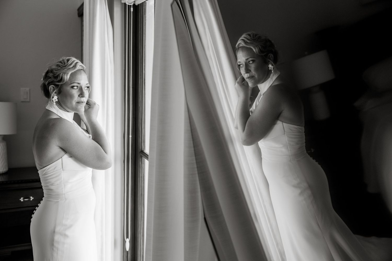 Black and white photo of a bride in a strapless gown adjusting her earring by a window with sheer curtains.