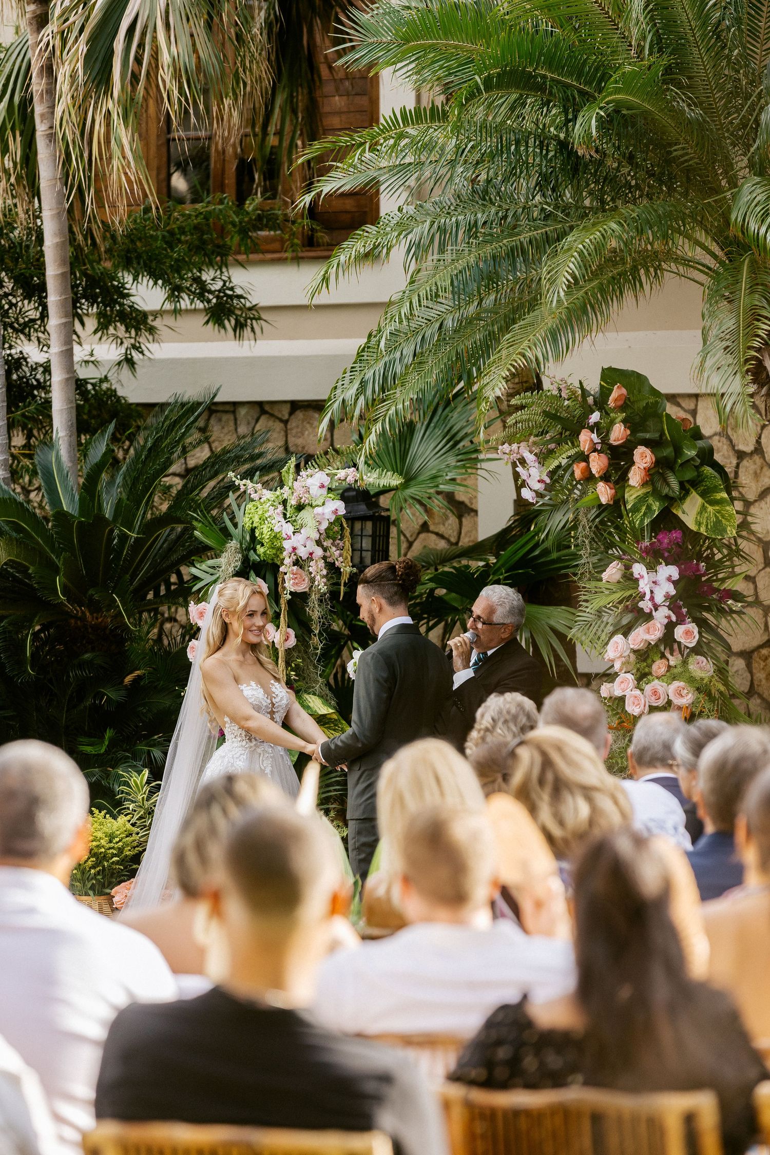 Outdoor tropical wedding ceremony with bride and groom at floral arch, guests seated, lush palm trees and pink flowers.