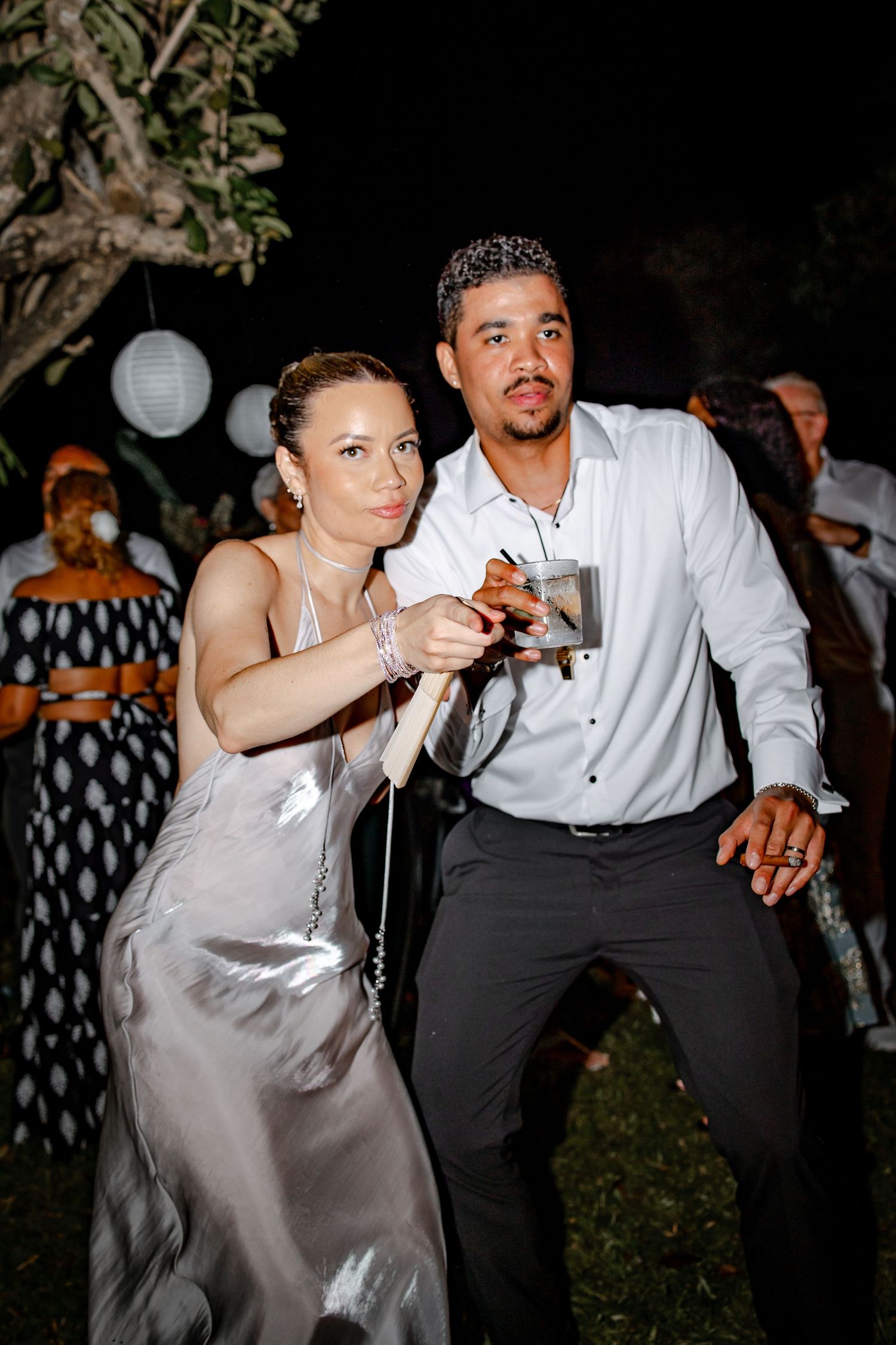 Couple dances together at an outdoor nighttime wedding reception, woman wearing a silver satin dress.
