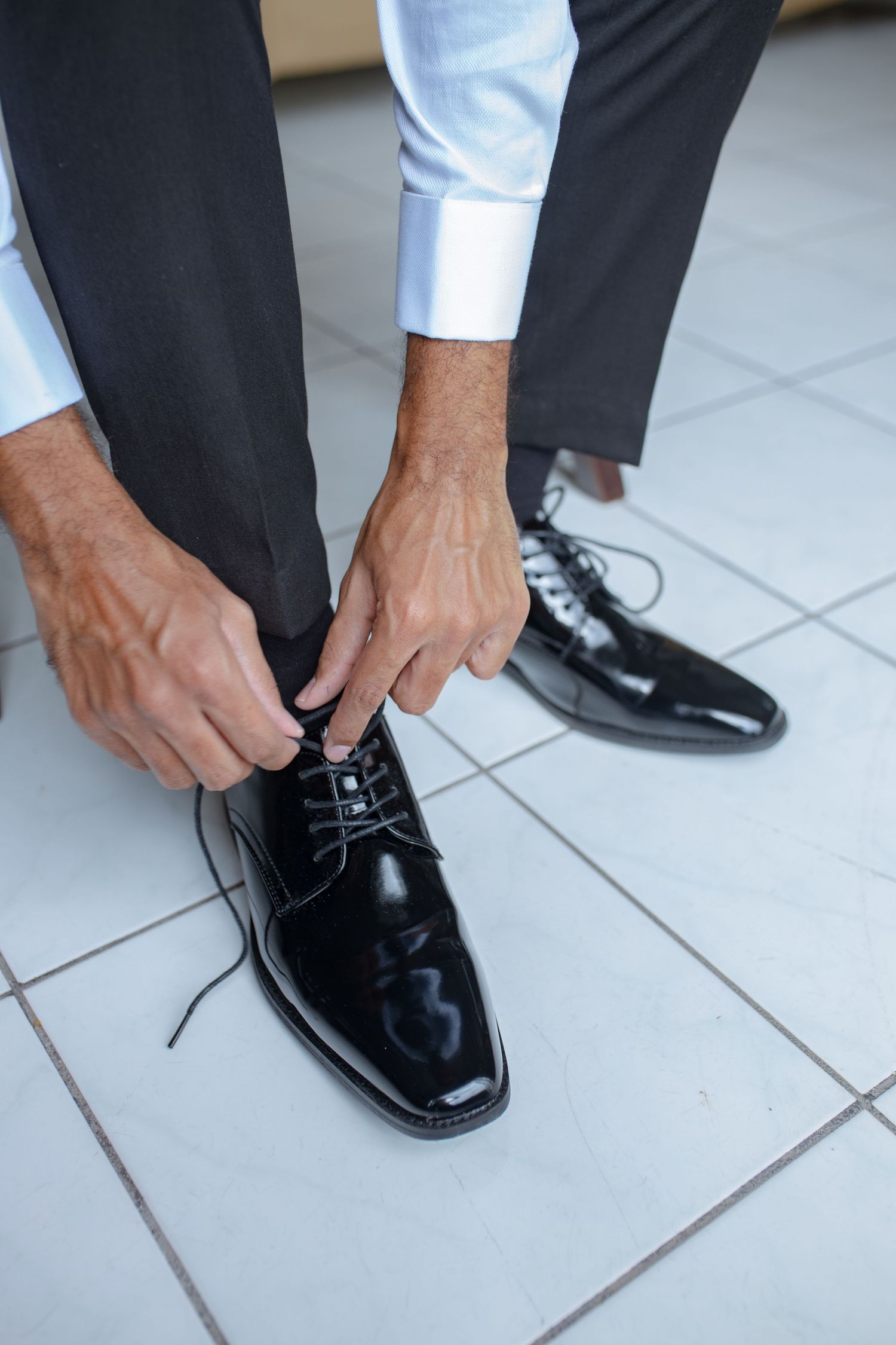 Man tying black patent leather dress shoes on tiled floor wearing formal suit.