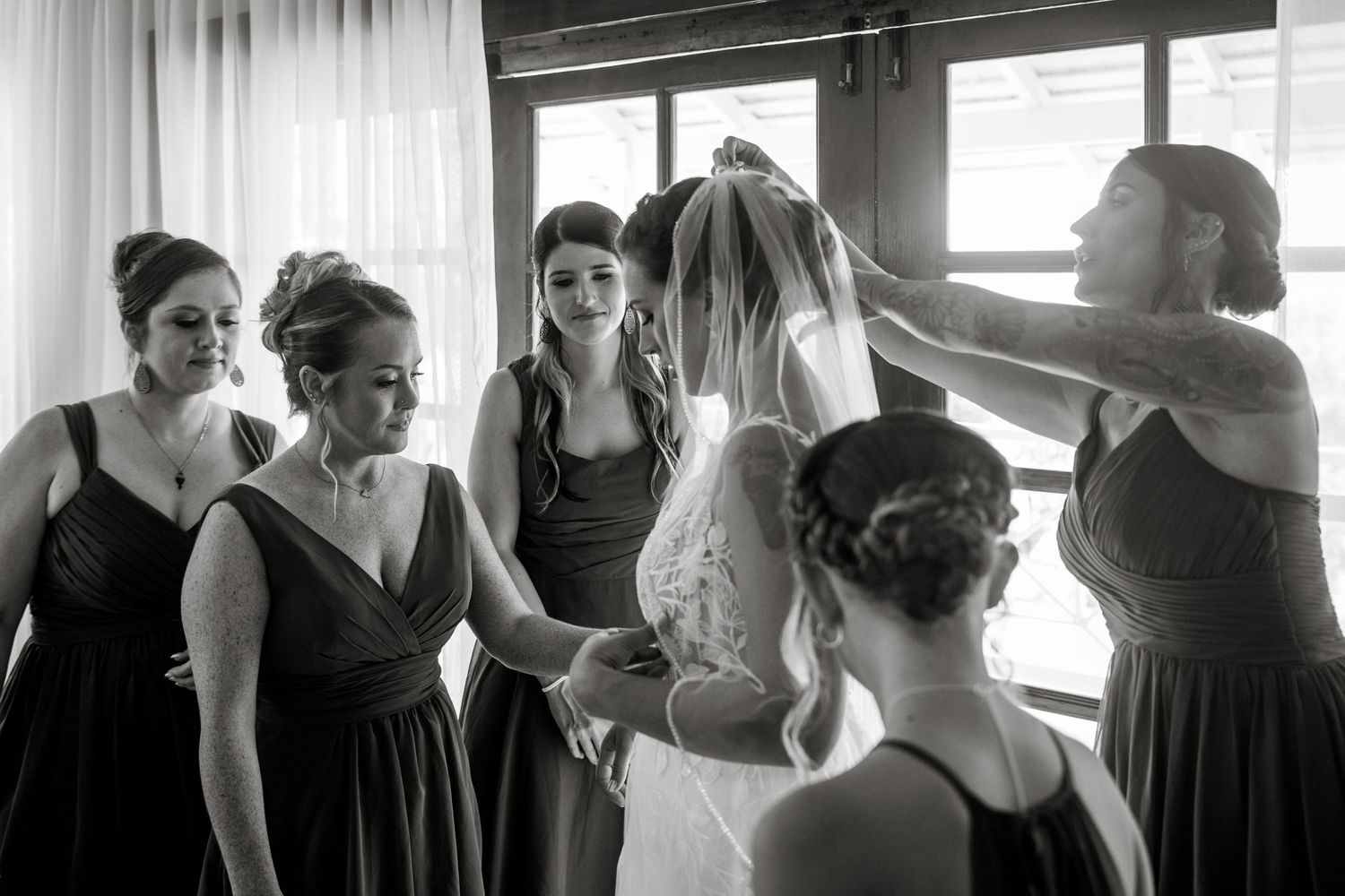 Black and white photo of bridesmaids placing veil on bride in elegant lace wedding dress.