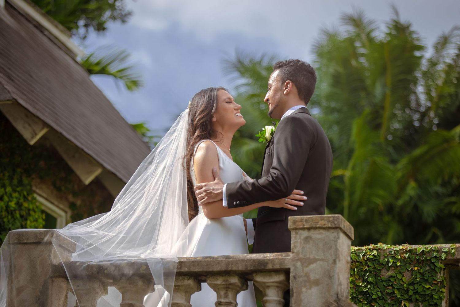 Bride and groom gazing at each other on a stone balcony surrounded by lush tropical greenery.