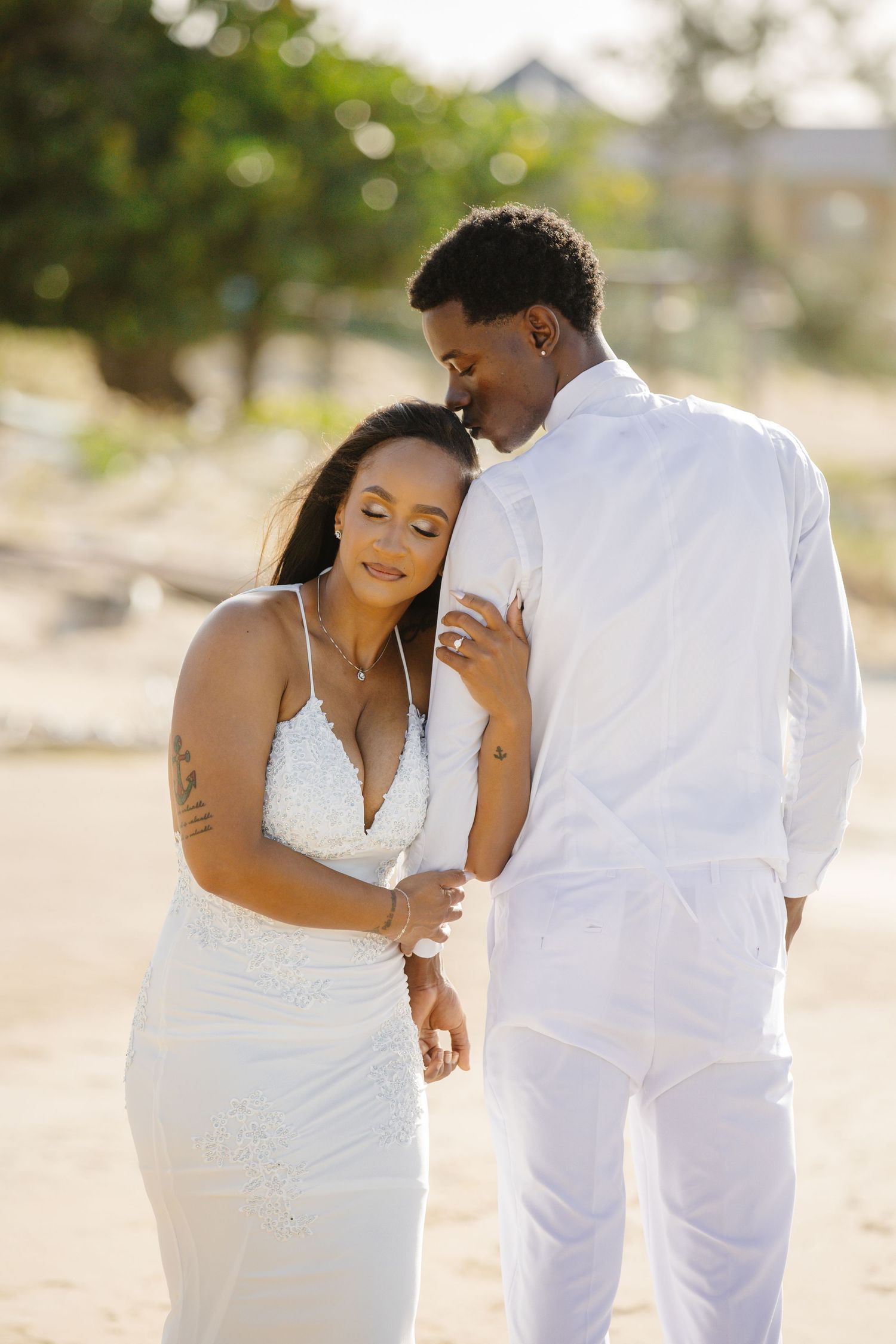 Black couple in white wedding attire share intimate moment on sunny sandy beach at golden hour.