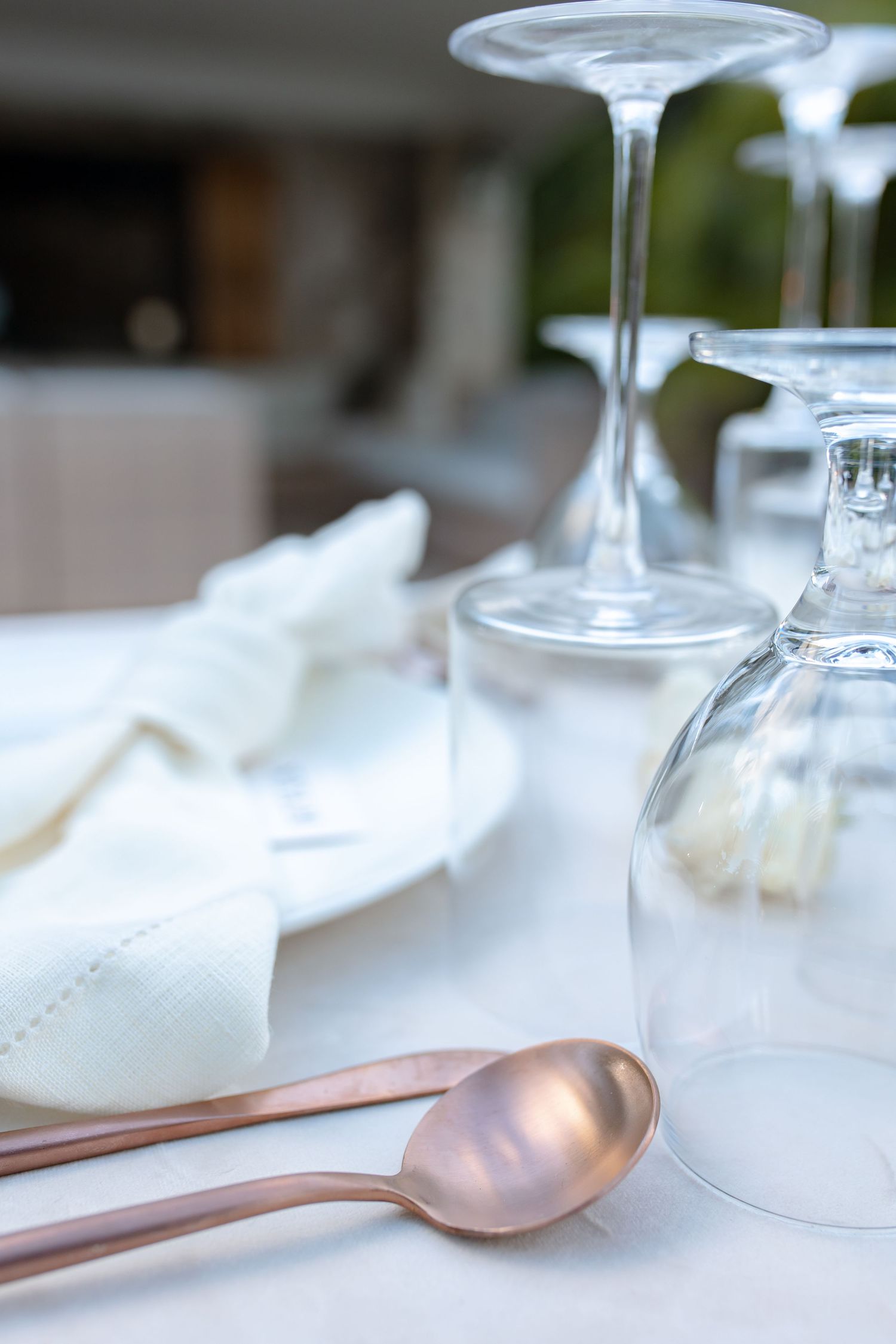 Elegant table setting with rose gold spoons, upside-down wine glasses, and a white linen napkin on a white tablecloth.