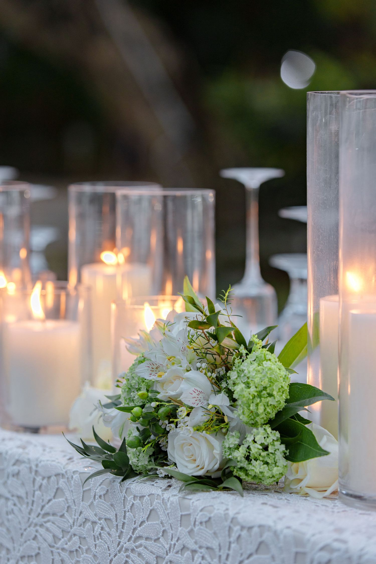 White roses and hydrangea bouquet surrounded by glowing candles on a lace-covered wedding table.