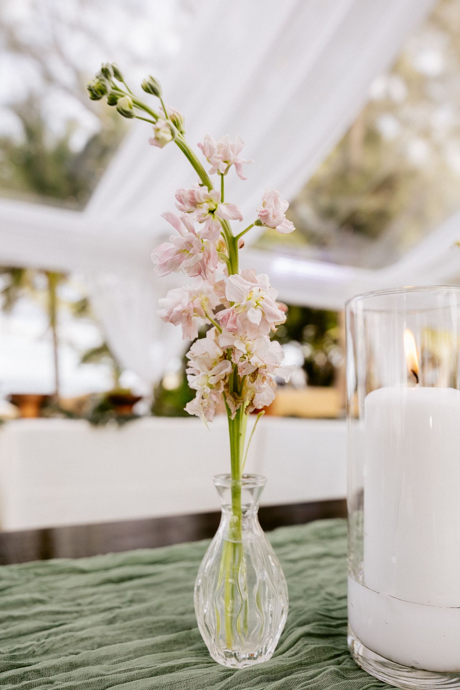 Delicate pink stock flowers in a glass bud vase beside a lit white pillar candle on a green tablecloth.