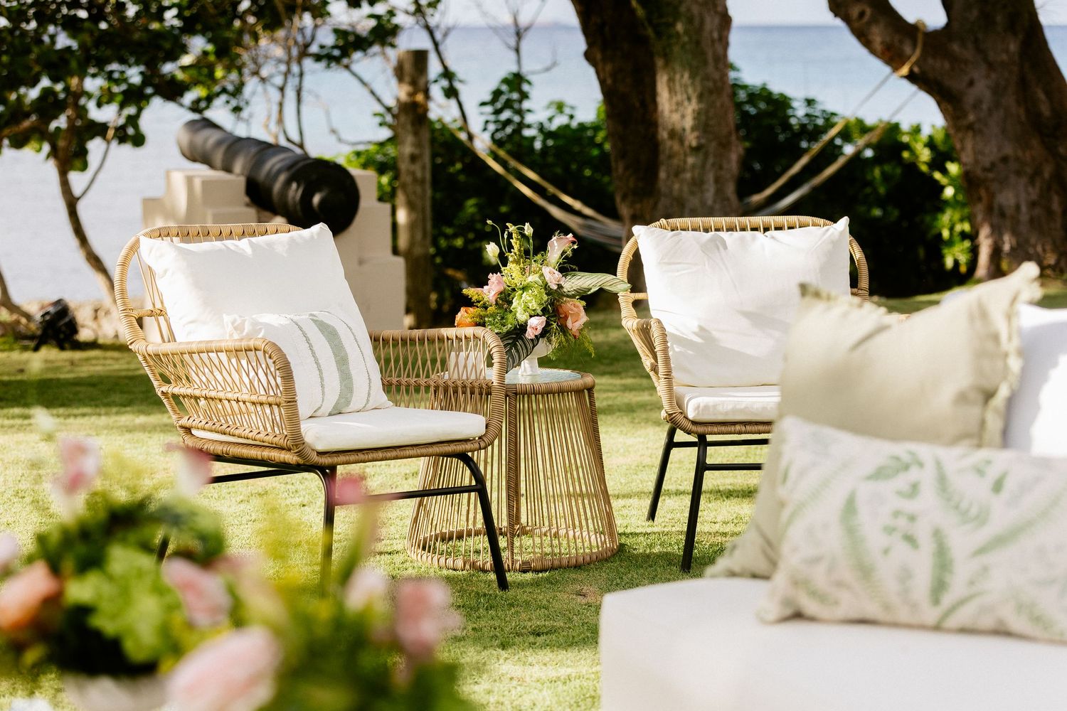 Elegant outdoor seating area with rattan chairs, white cushions, and floral centerpiece on a lush green lawn near the ocean.