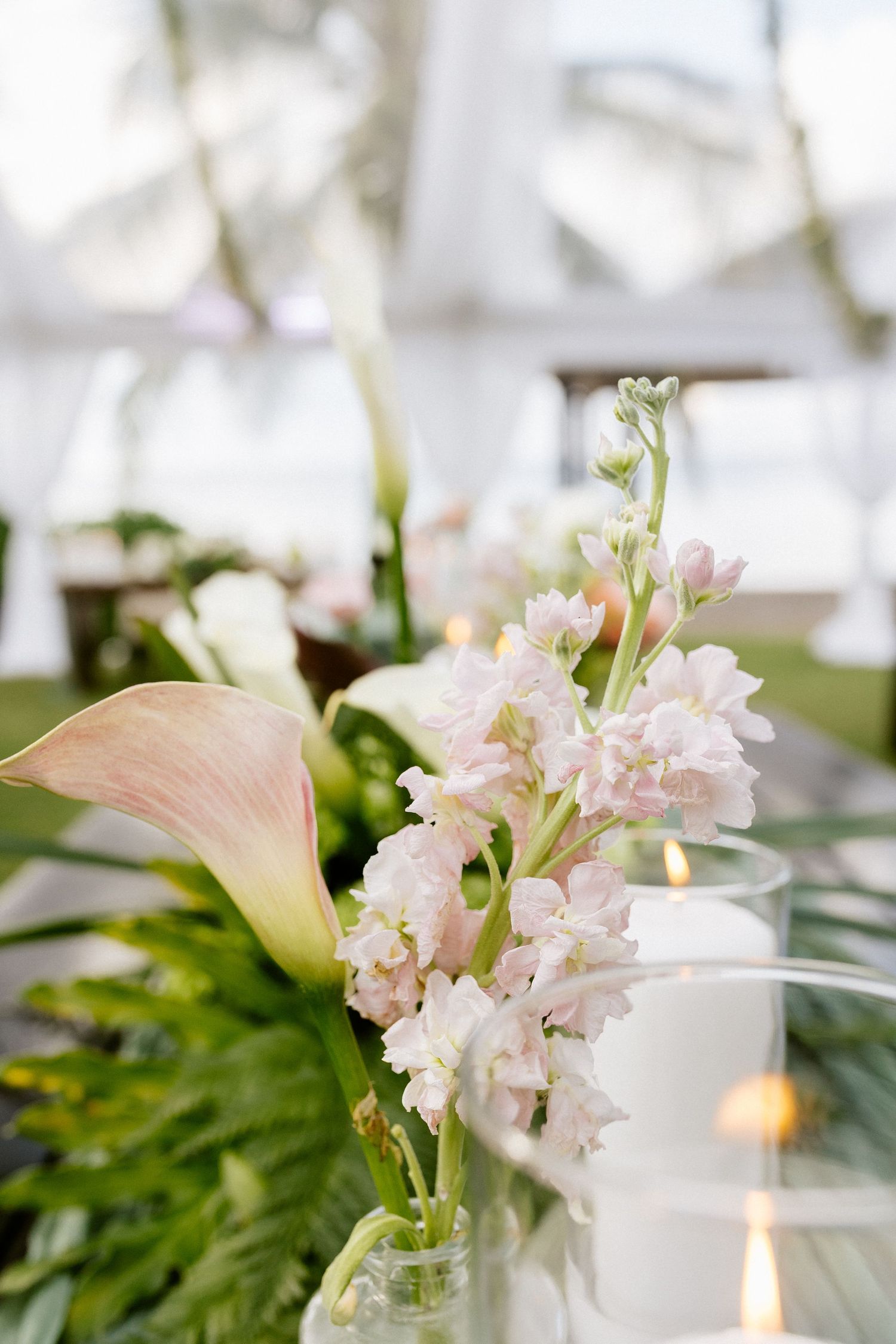 Elegant wedding table centerpiece with blush pink calla lilies, white stock flowers, tropical greenery, and glowing candles.