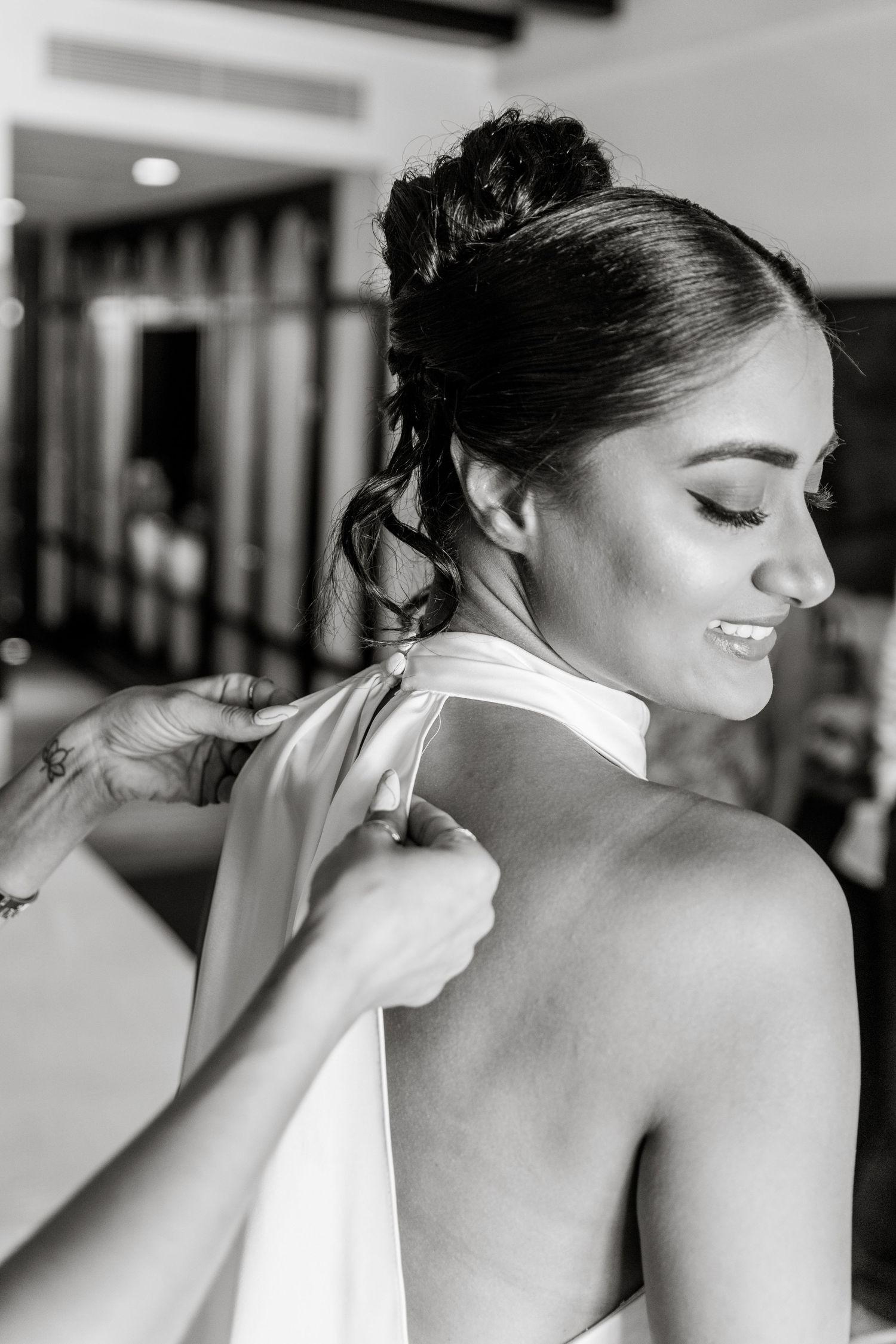 Black and white photo of a smiling bride having her halter wedding dress tied by a tattooed assistant.