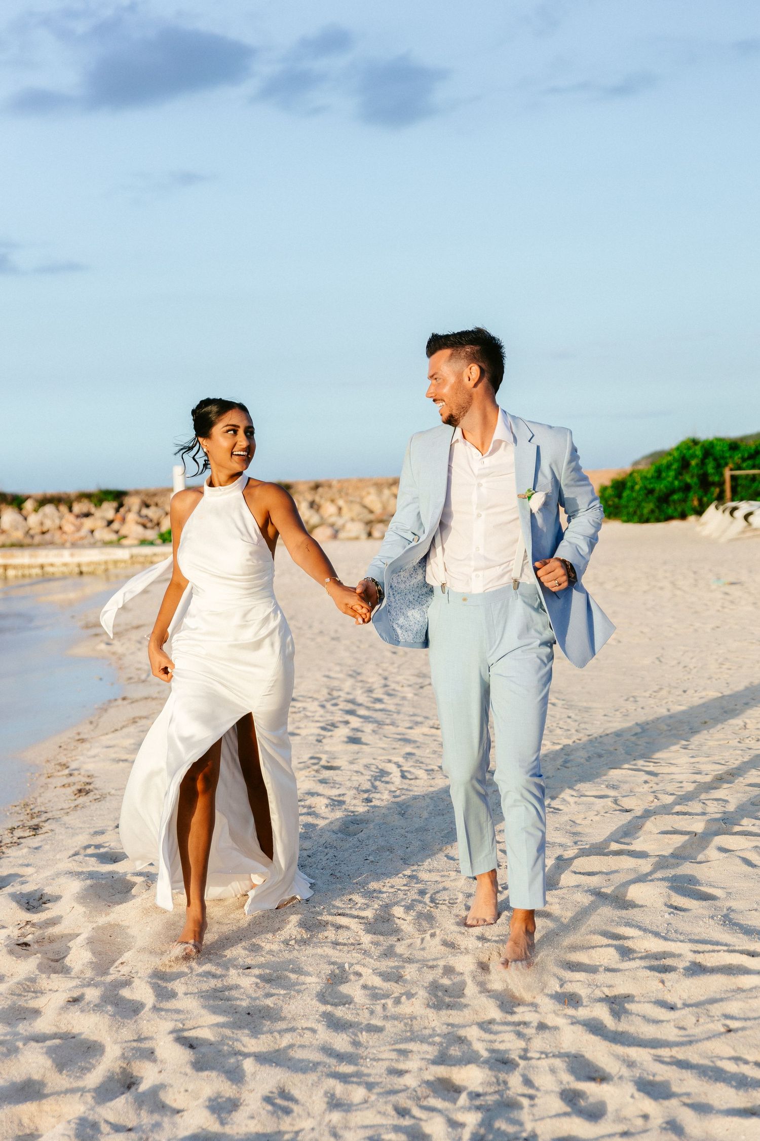 Newlywed couple holding hands running barefoot on sandy beach at sunset after destination wedding.