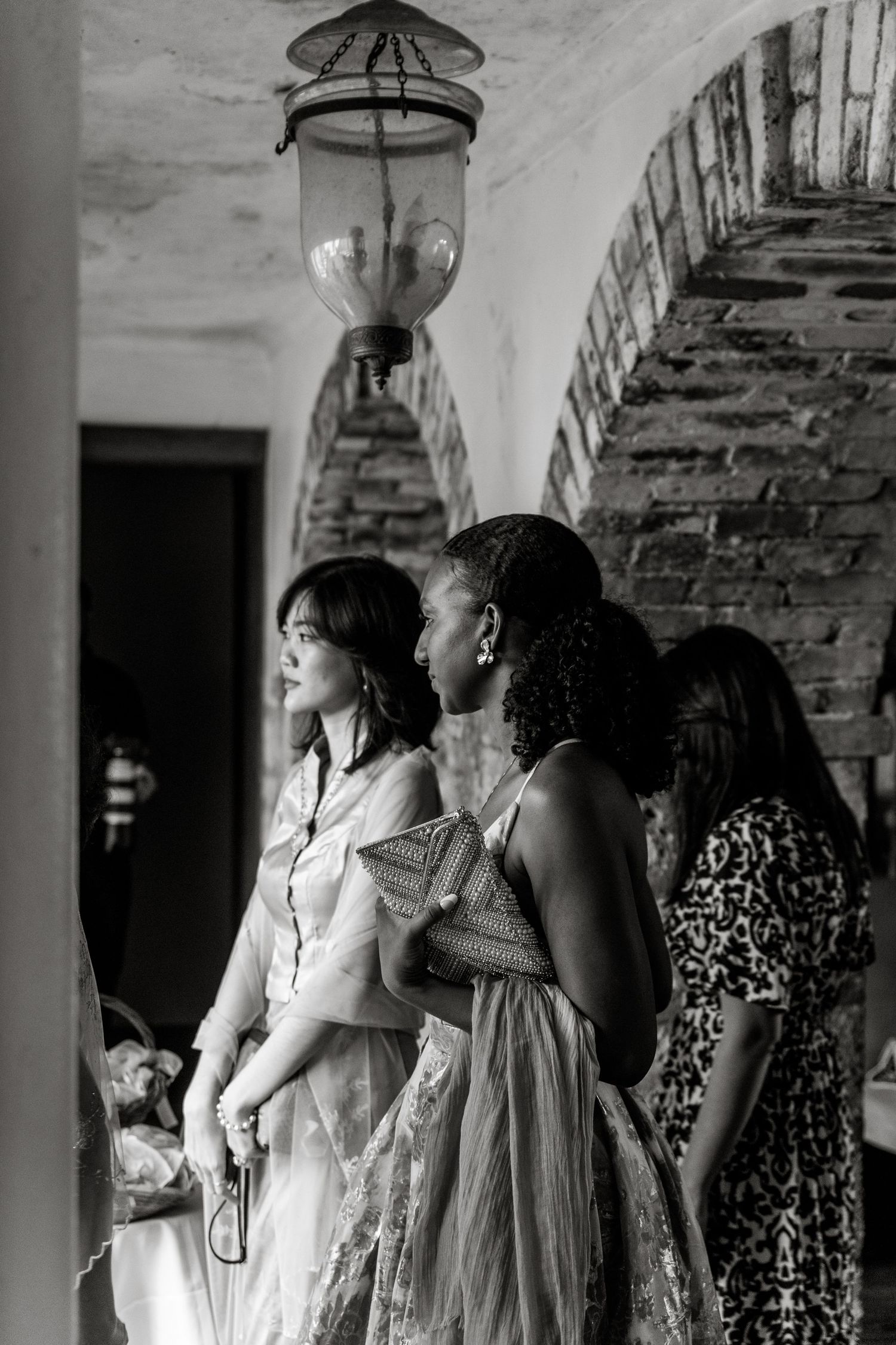 Black and white photo of elegantly dressed women standing under stone arch with hanging lantern at wedding.