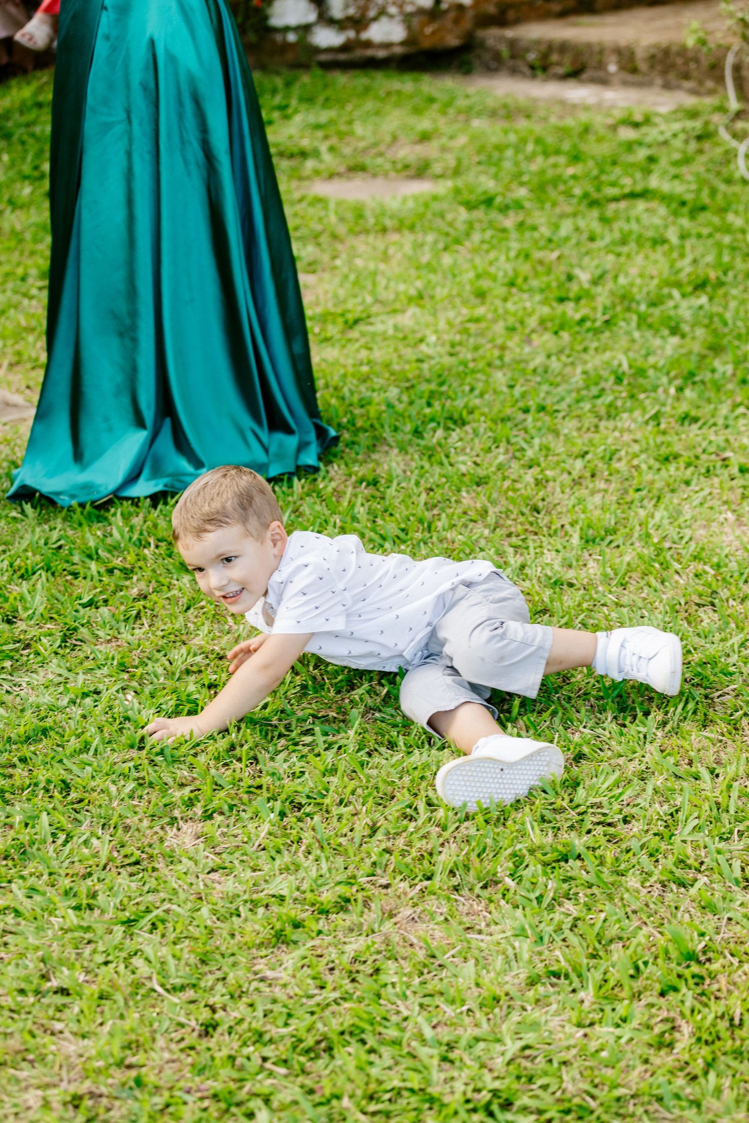 Young boy in white shirt and gray shorts playing on green grass at outdoor wedding.