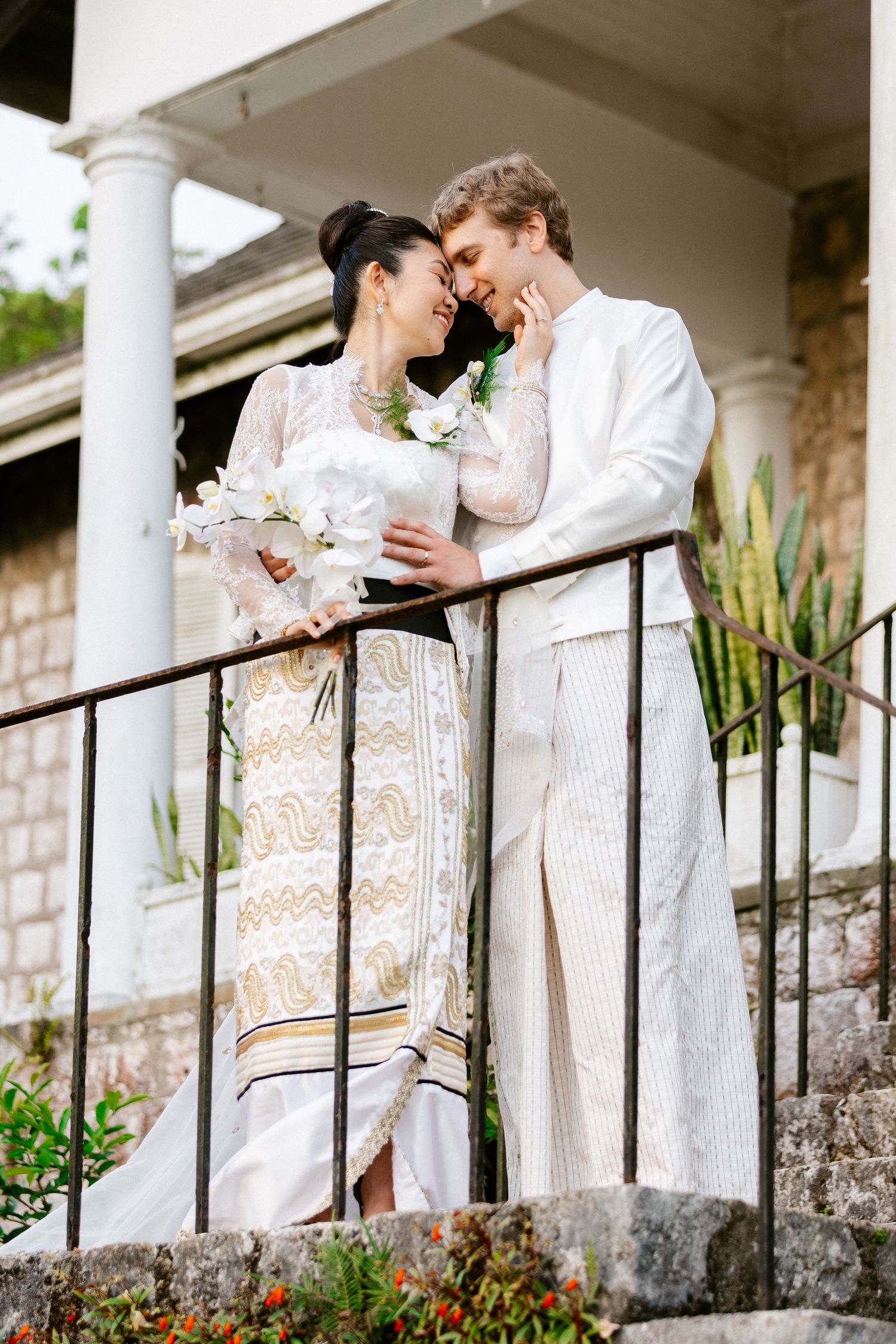 Bride and groom in traditional Thai wedding attire sharing romantic moment on stone balcony.