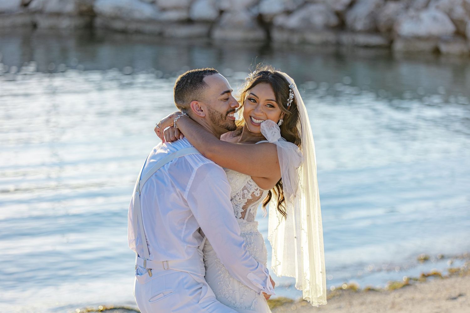 Newlyweds laughing and embracing on a sunny beach shoreline in white wedding attire.