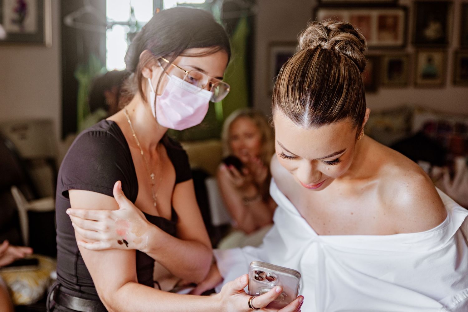 Masked makeup artist showing bride details on phone during bridal preparation at home.