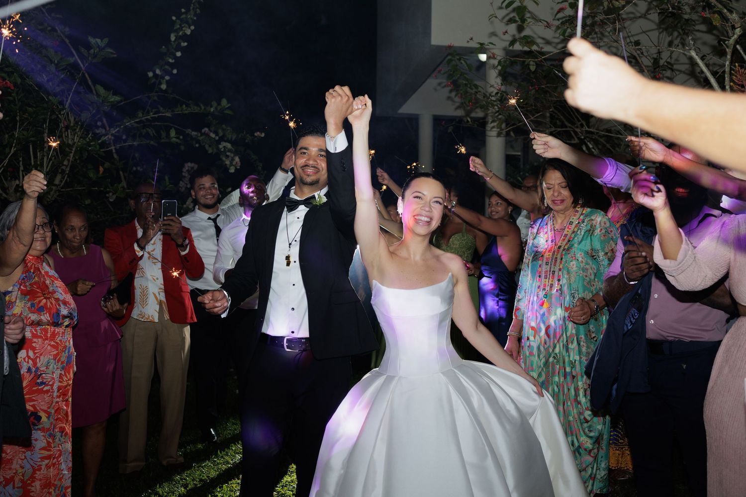 Newlyweds celebrate joyfully with raised hands as guests wave sparklers during outdoor evening wedding reception.