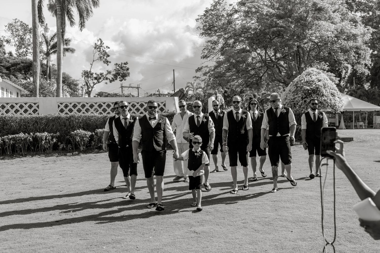 Black and white photo of groomsmen in matching vests walking together at outdoor wedding venue.