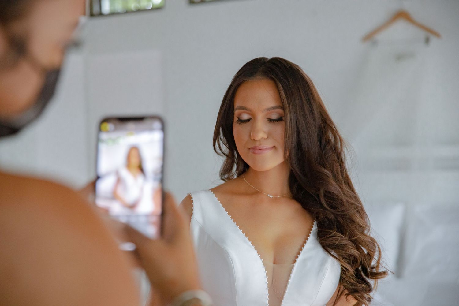 Groom photographing bride in white wedding gown with deep V-neckline on smartphone.