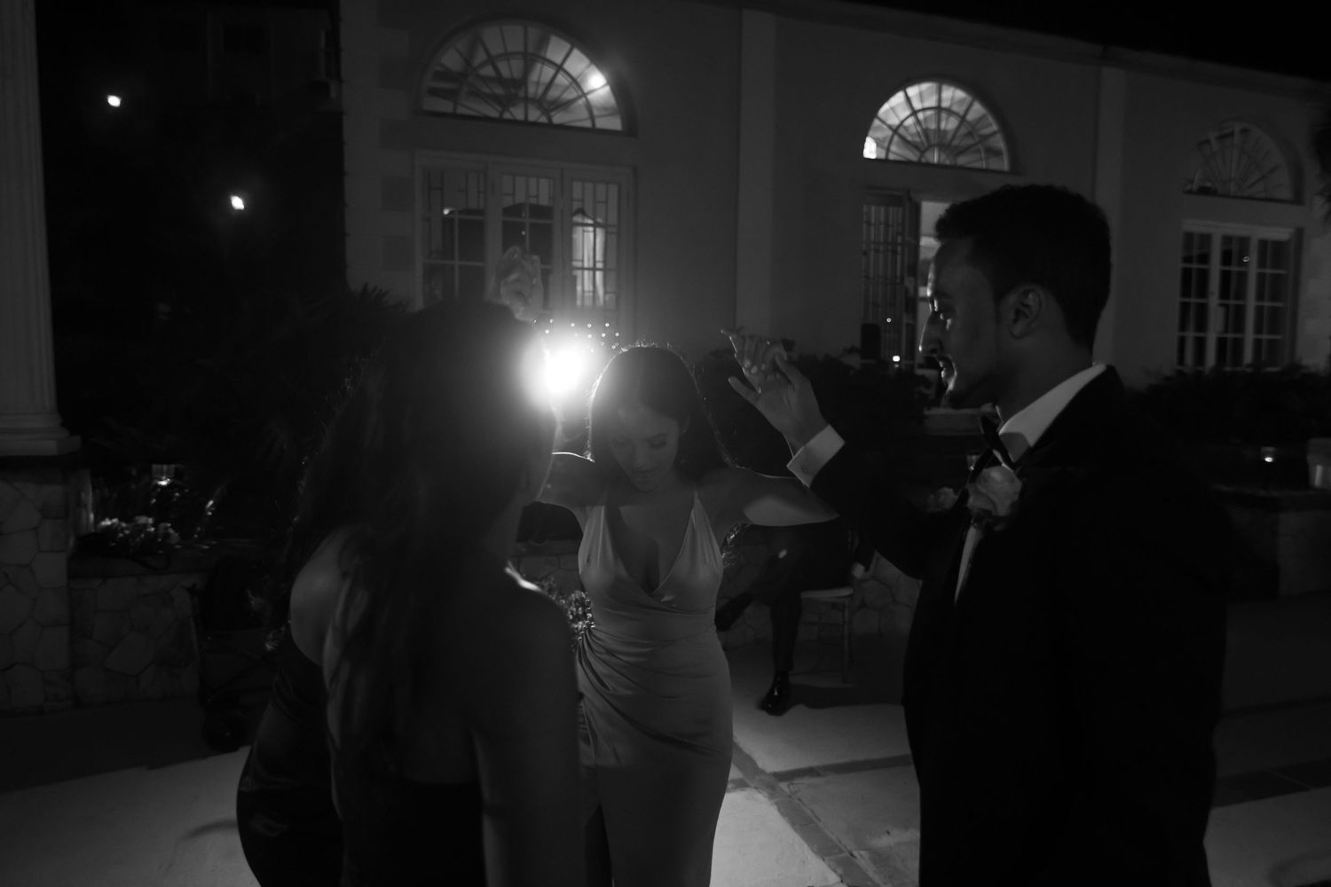 Black and white photo of wedding guests celebrating with sparklers outside an illuminated venue at night.