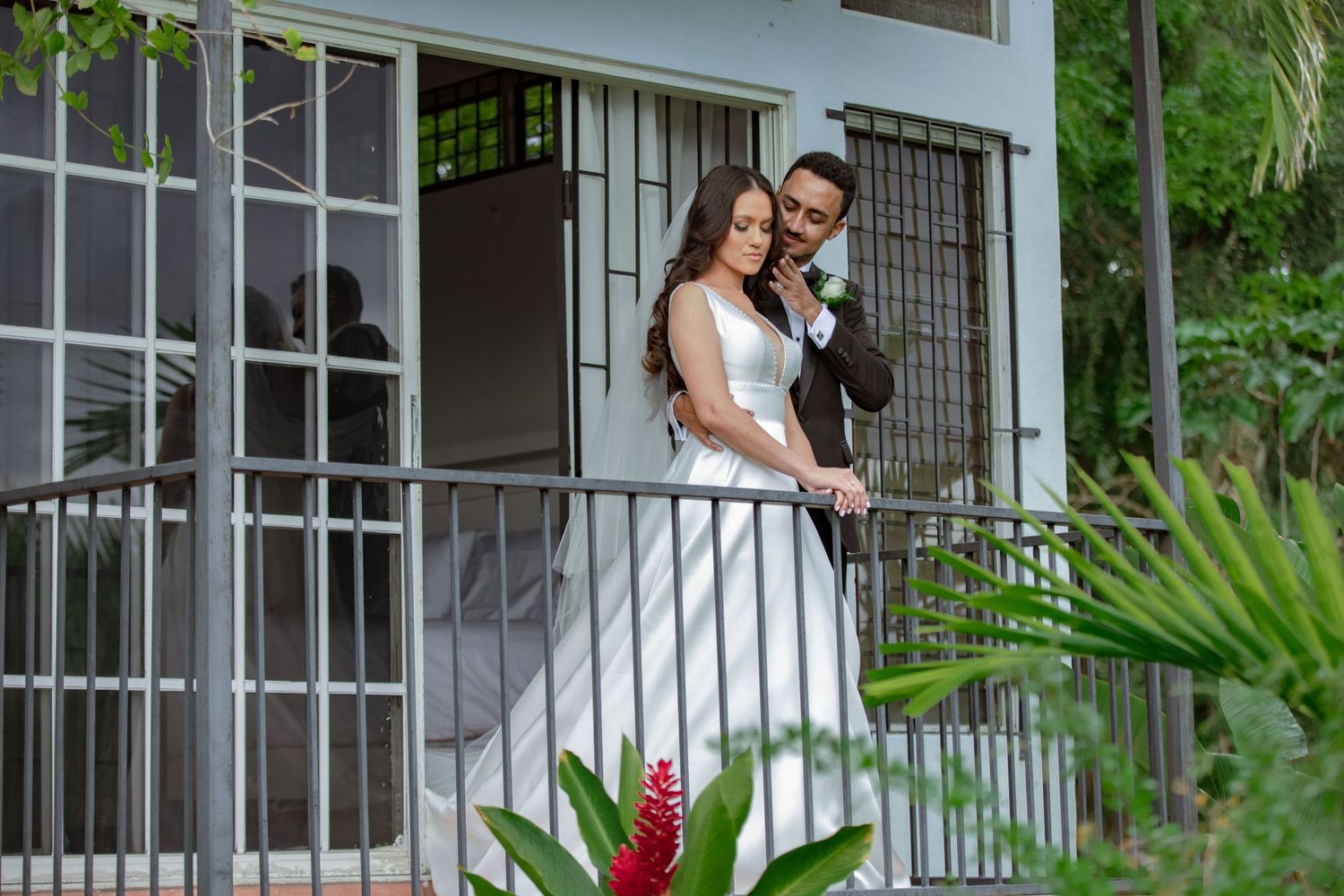 Bride and groom share intimate moment on white building balcony surrounded by tropical plants.