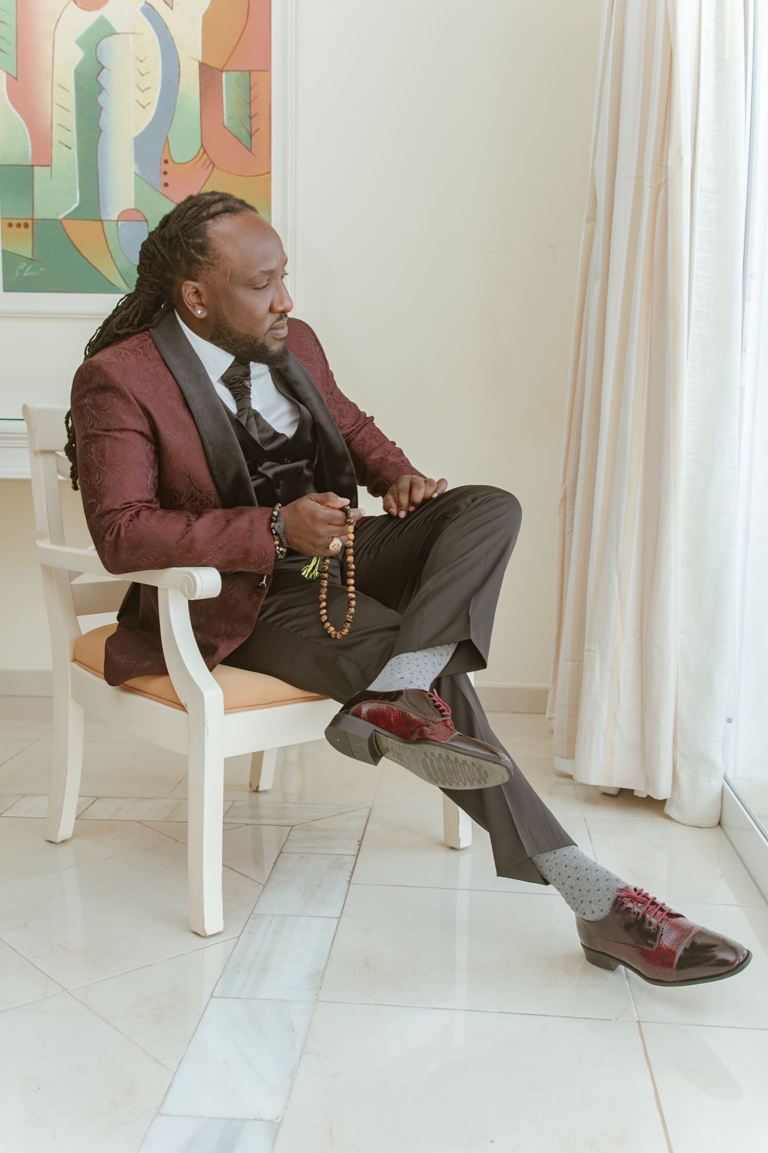 Groom seated in white chair wearing burgundy tuxedo, holding prayer beads, looking contemplative.